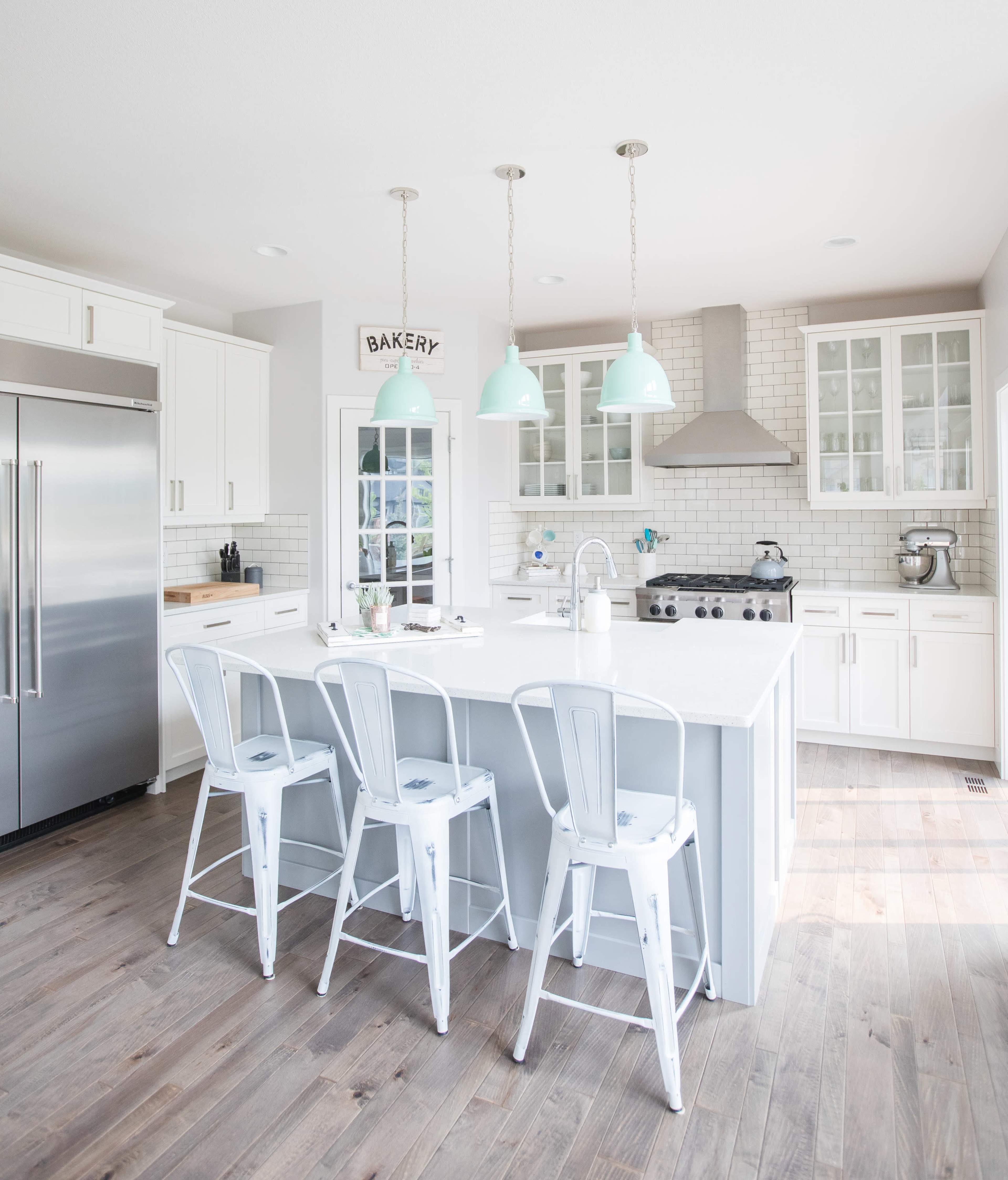 A modern kitchen features white cabinets, stainless steel appliances, a large island with seating, and pendant lights hanging above.