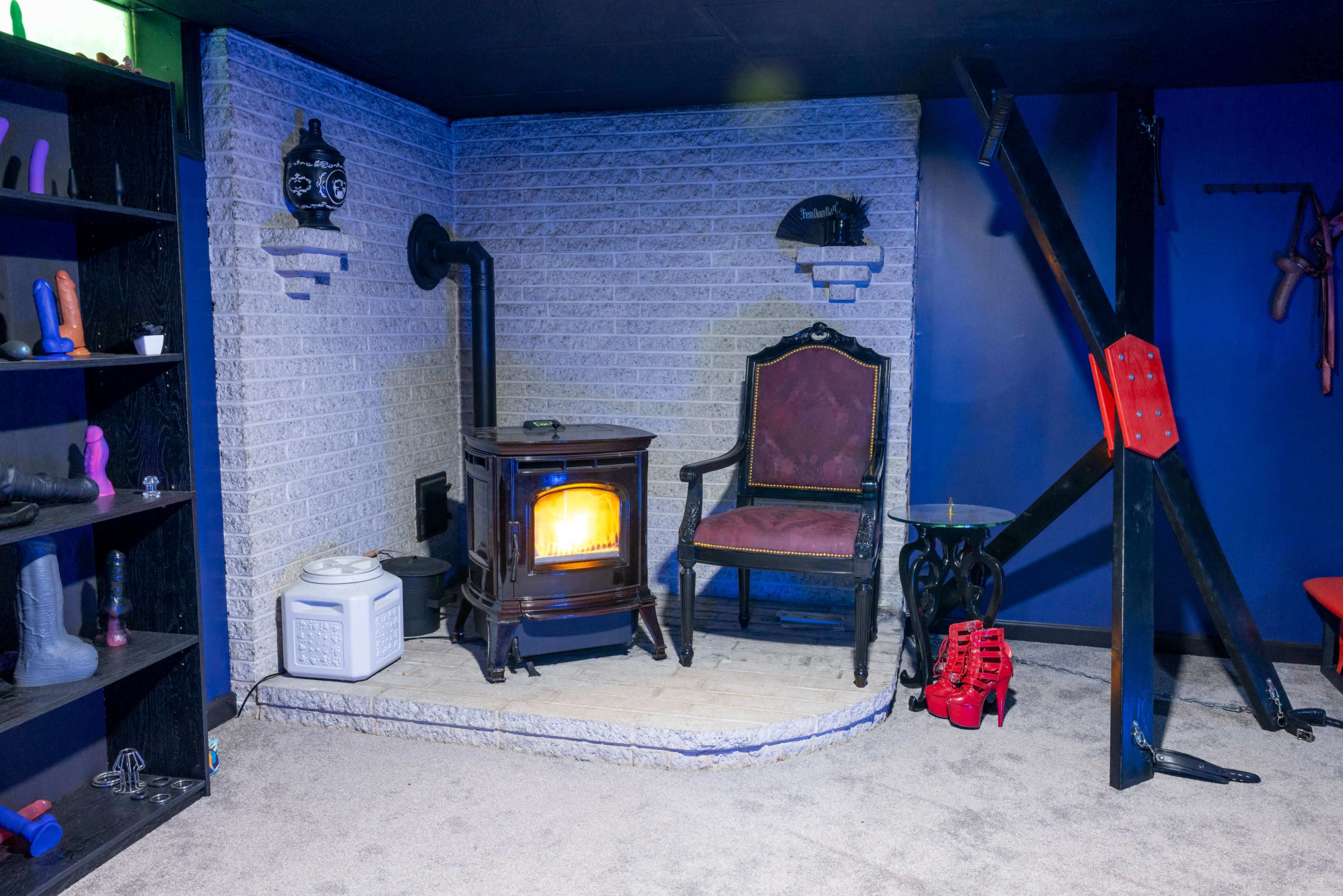 The image shows a dimly lit room featuring a stove, a pair of chairs, and various items on shelves, with a distinctive cross structure against the wall.