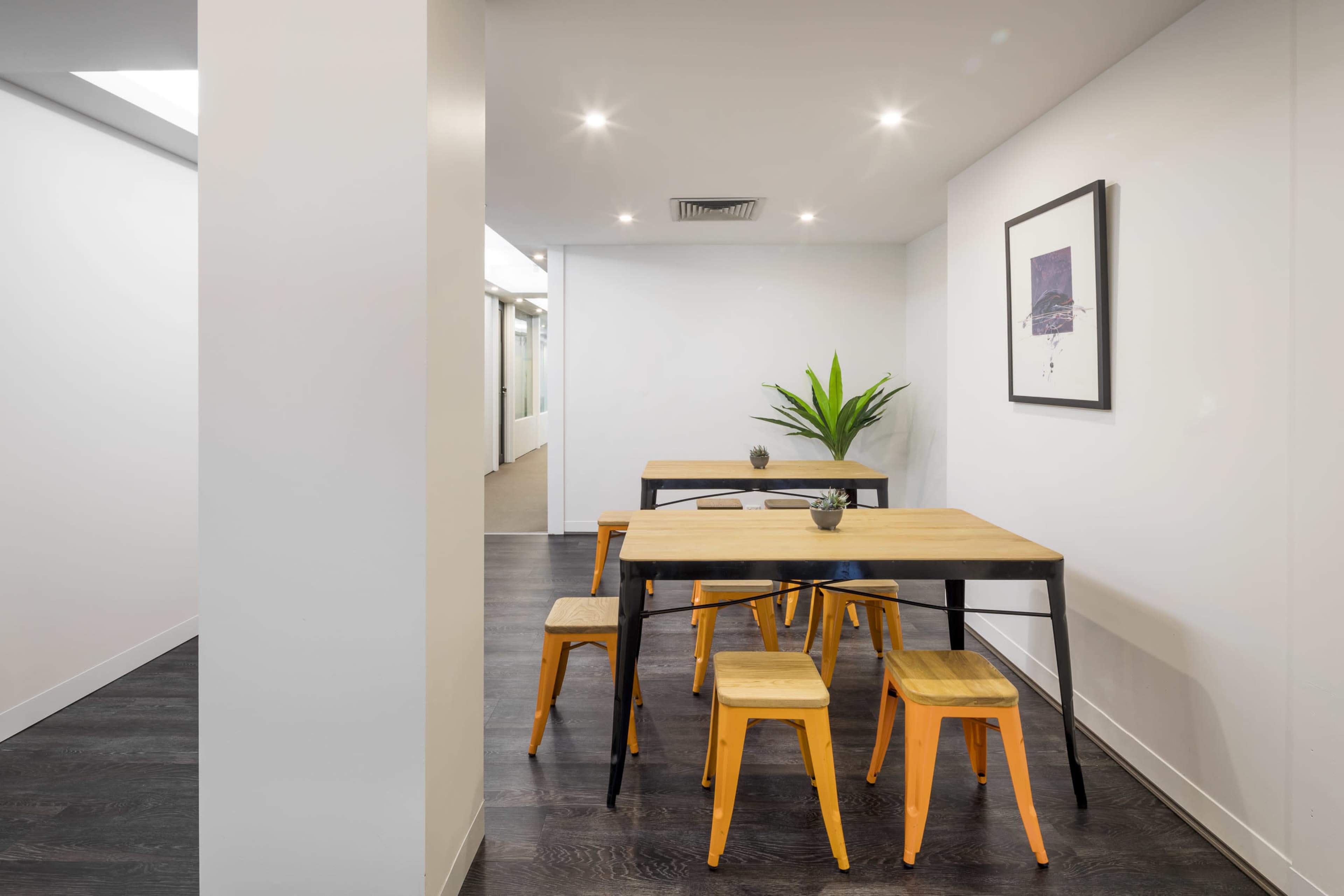 A modern interior features a wooden table with black legs surrounded by four orange-accented stools, positioned next to a potted plant against a white wall.