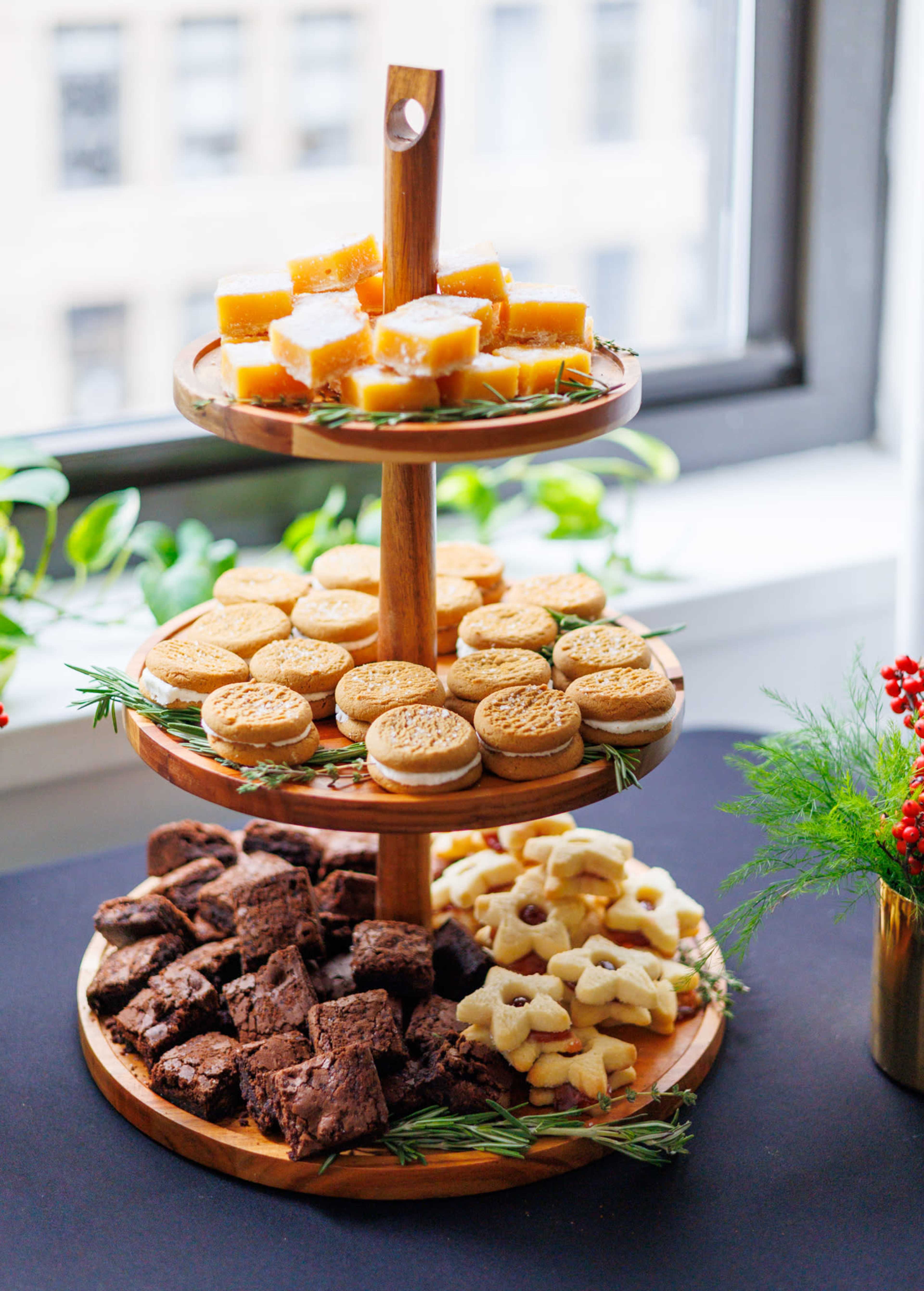 A three-tiered wooden stand displays an assortment of desserts, including brownies, cookies, and yellow cakes, arranged with greenery.