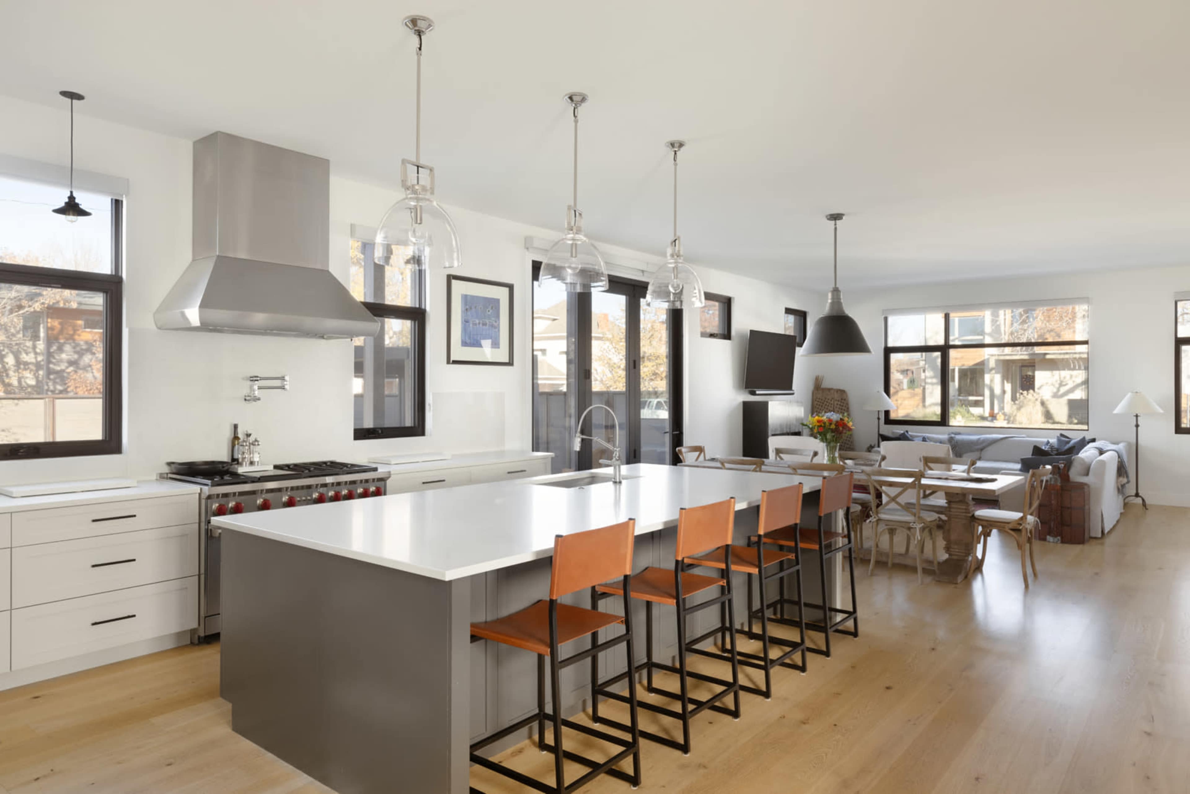 The image shows a modern kitchen with a large island, stainless steel appliances, and a dining area next to living space featuring large windows.