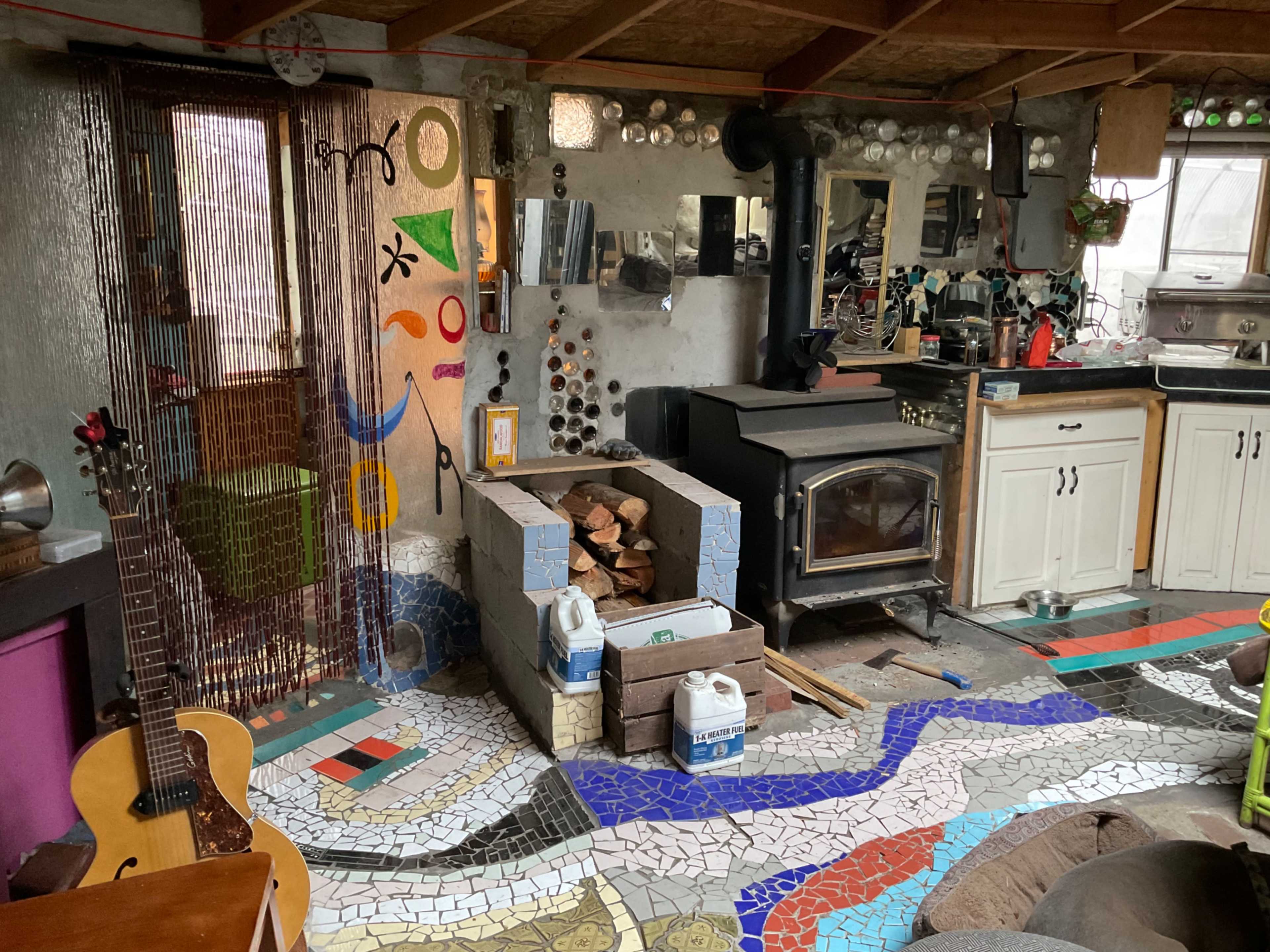 The image shows a cozy interior space featuring a wood-burning stove, a mosaic floor, a wooden guitar, and a kitchen area with cabinets and various utensils.