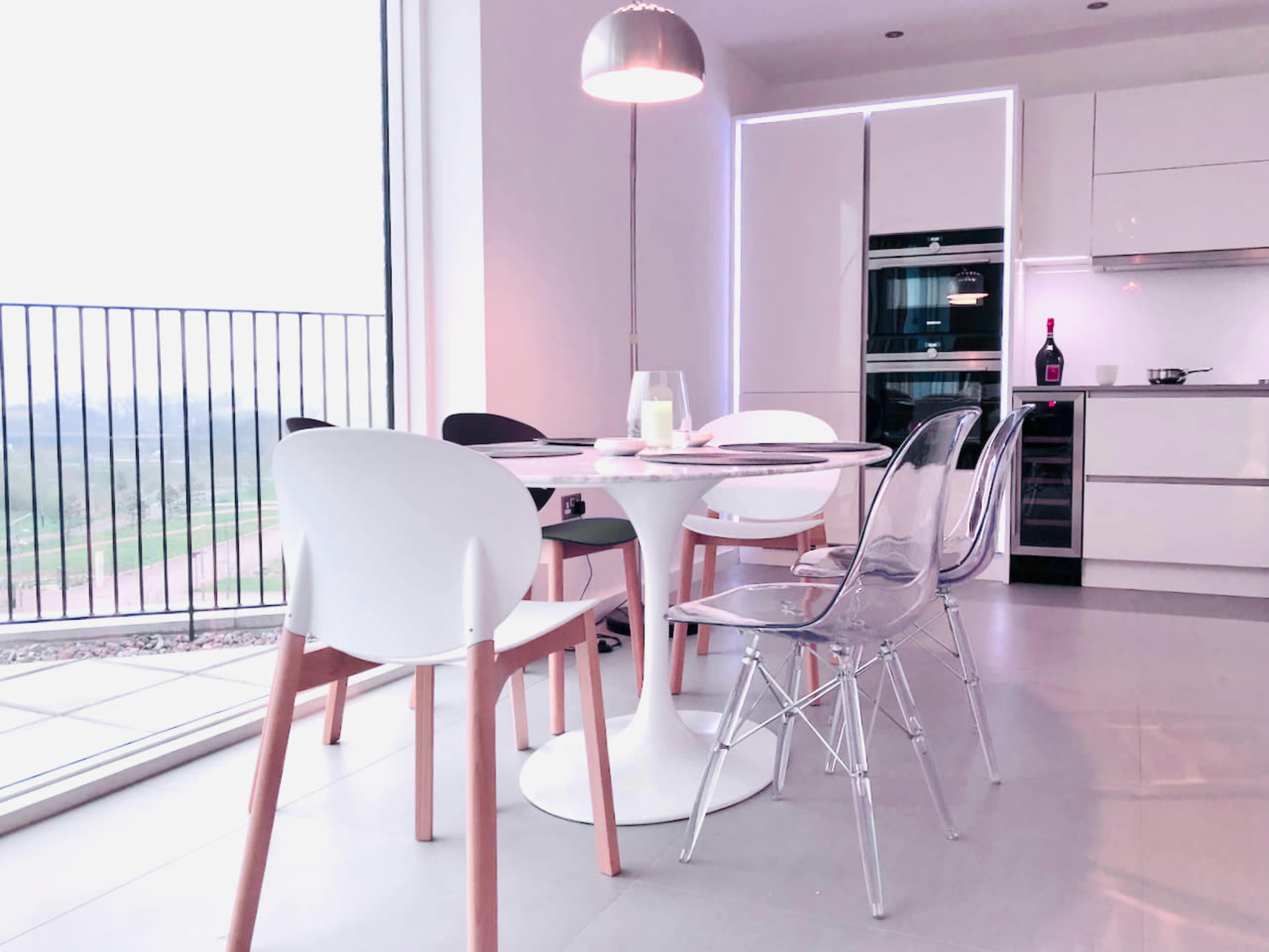 A modern dining area features a round table surrounded by white and clear chairs, with large windows offering a view of the landscape.