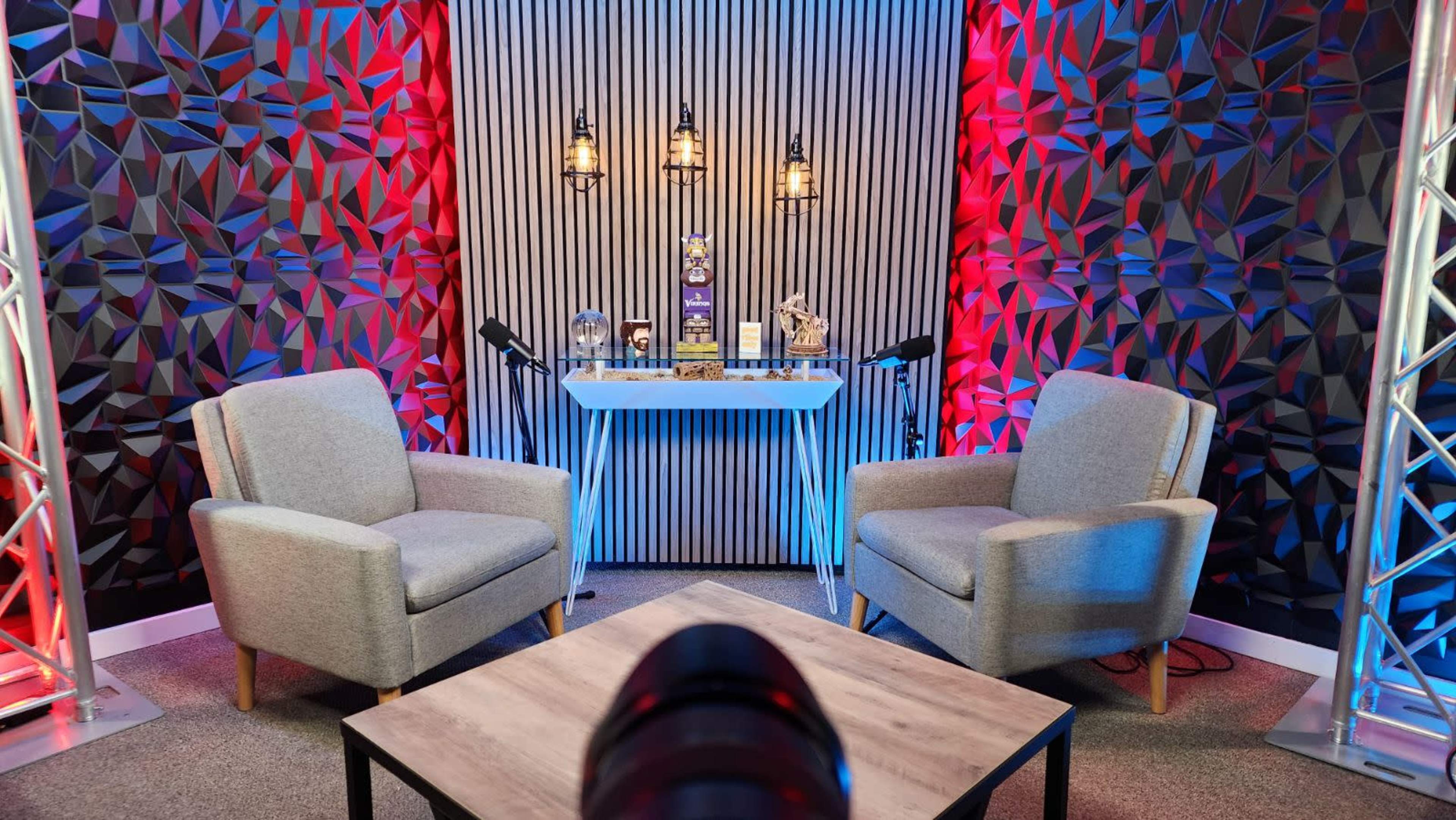 A modern podcasting studio featuring two gray chairs, a wooden table, and a backdrop with patterned walls illuminated by red and blue lights.