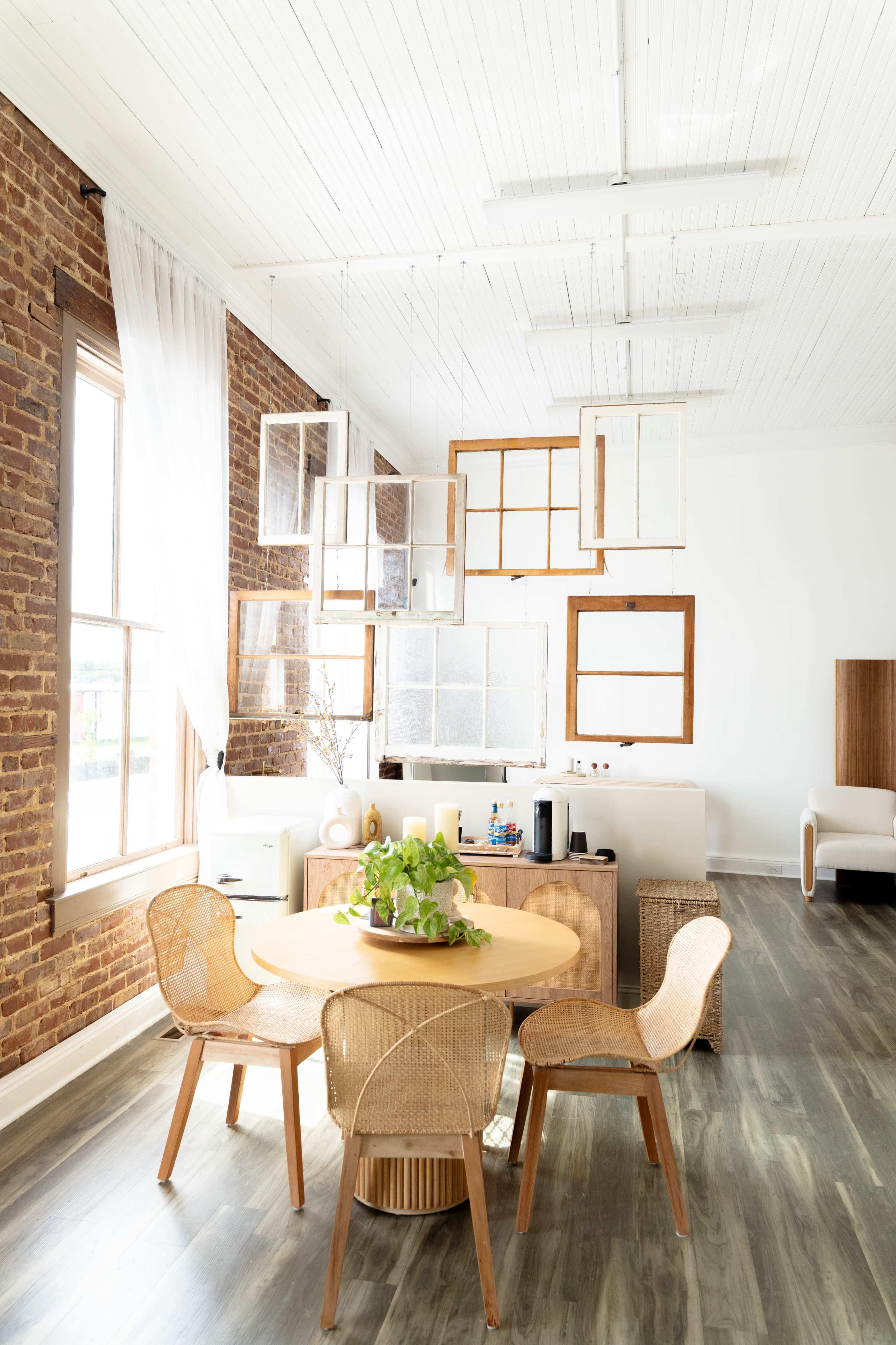 A round wooden dining table with four woven chairs is positioned in a bright room featuring exposed brick walls and large windows.