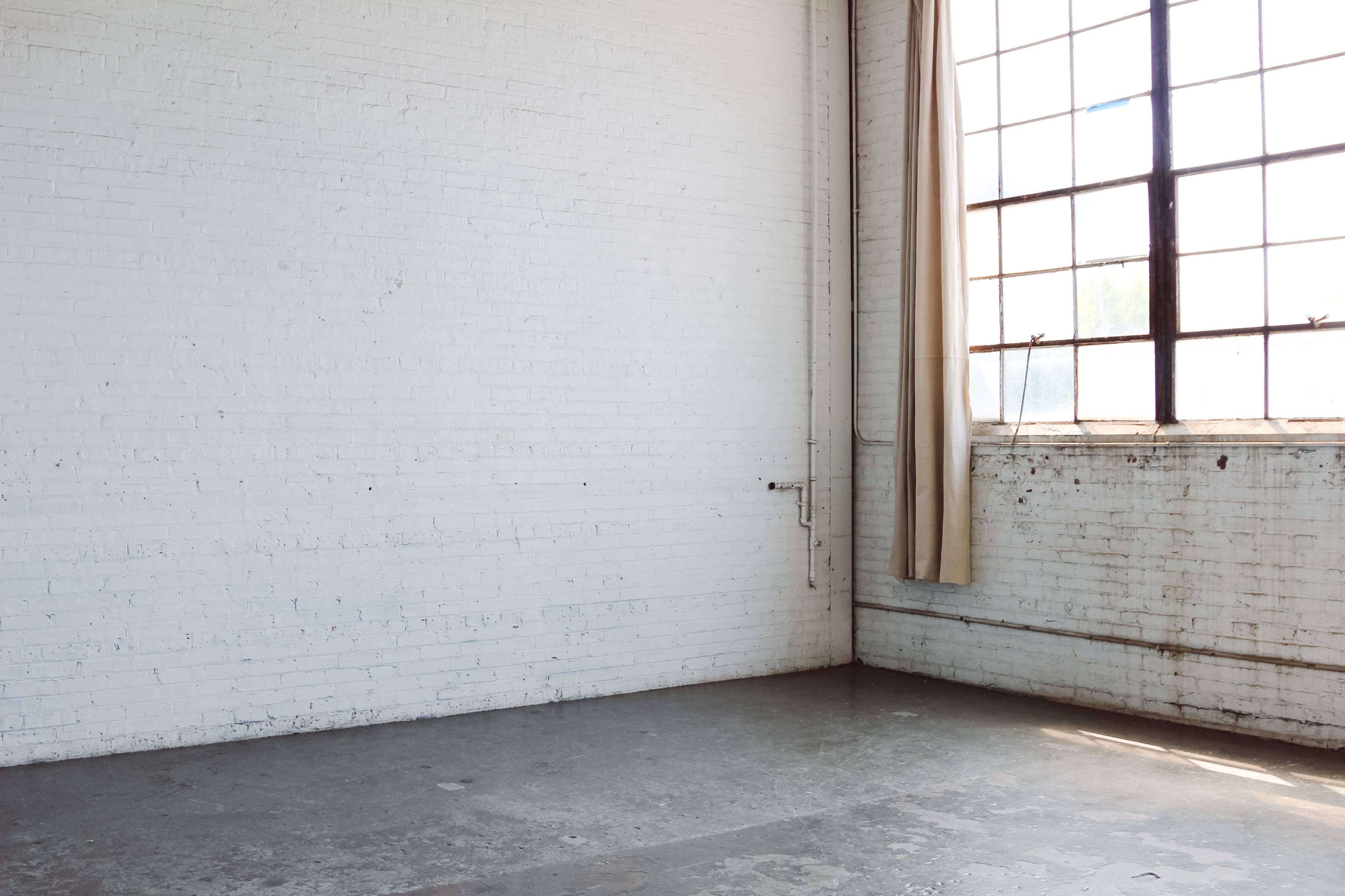 A vacant room with white brick walls, a large window letting in natural light, and a single curtain draped beside the window.