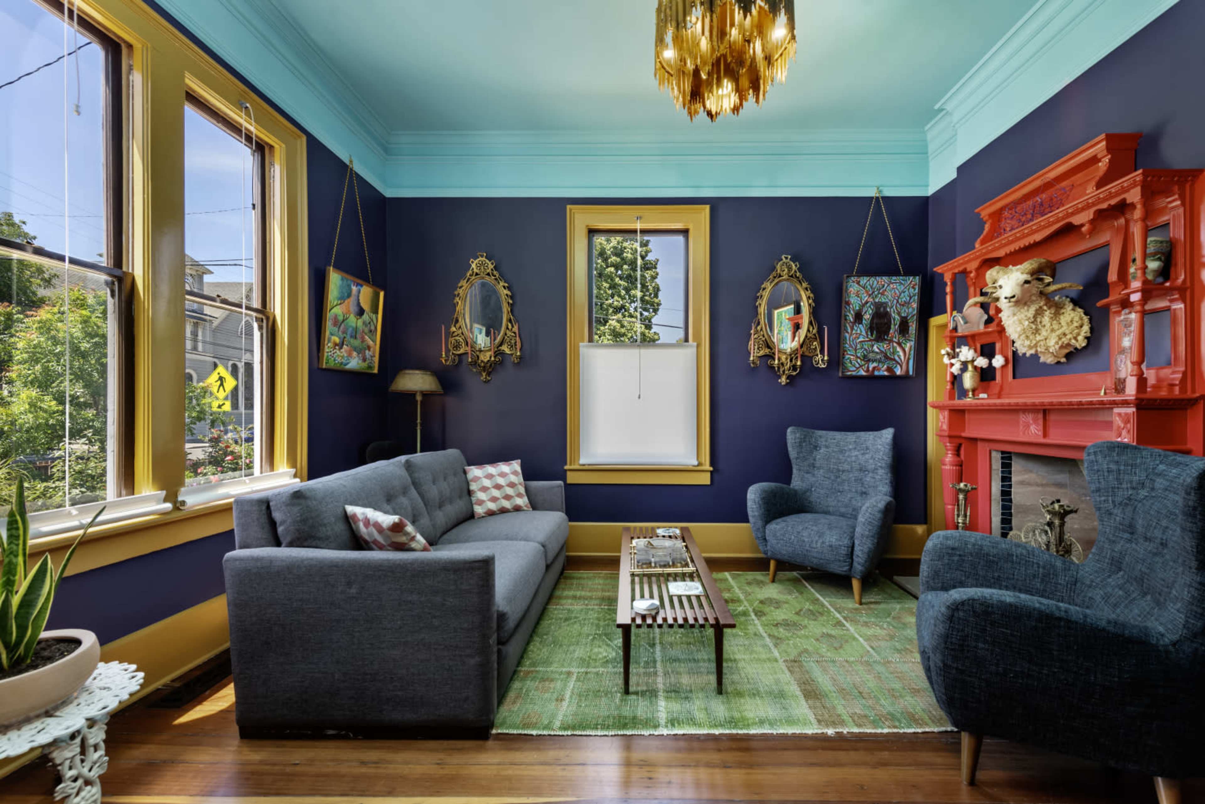 The room features a blue and teal color scheme, with a dark blue wall, large windows, a gray couch, and a bright red mantelpiece adorned with decorative items.