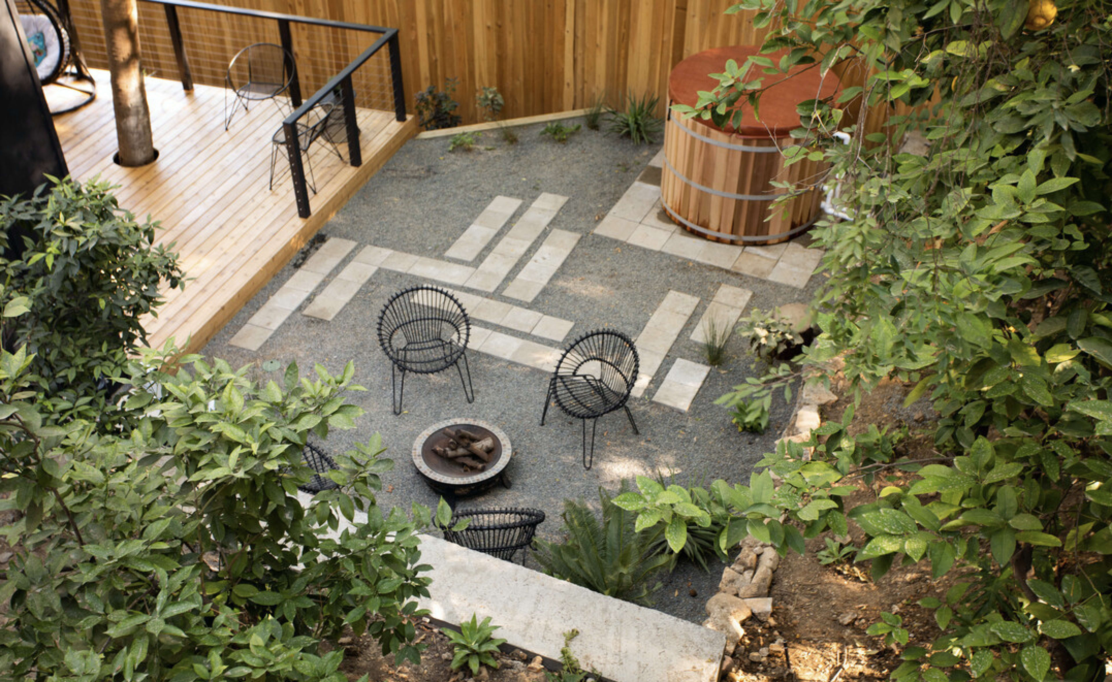 A landscaped outdoor area with gravel pathways, a fire pit surrounded by chairs, and a hot tub, framed by greenery.
