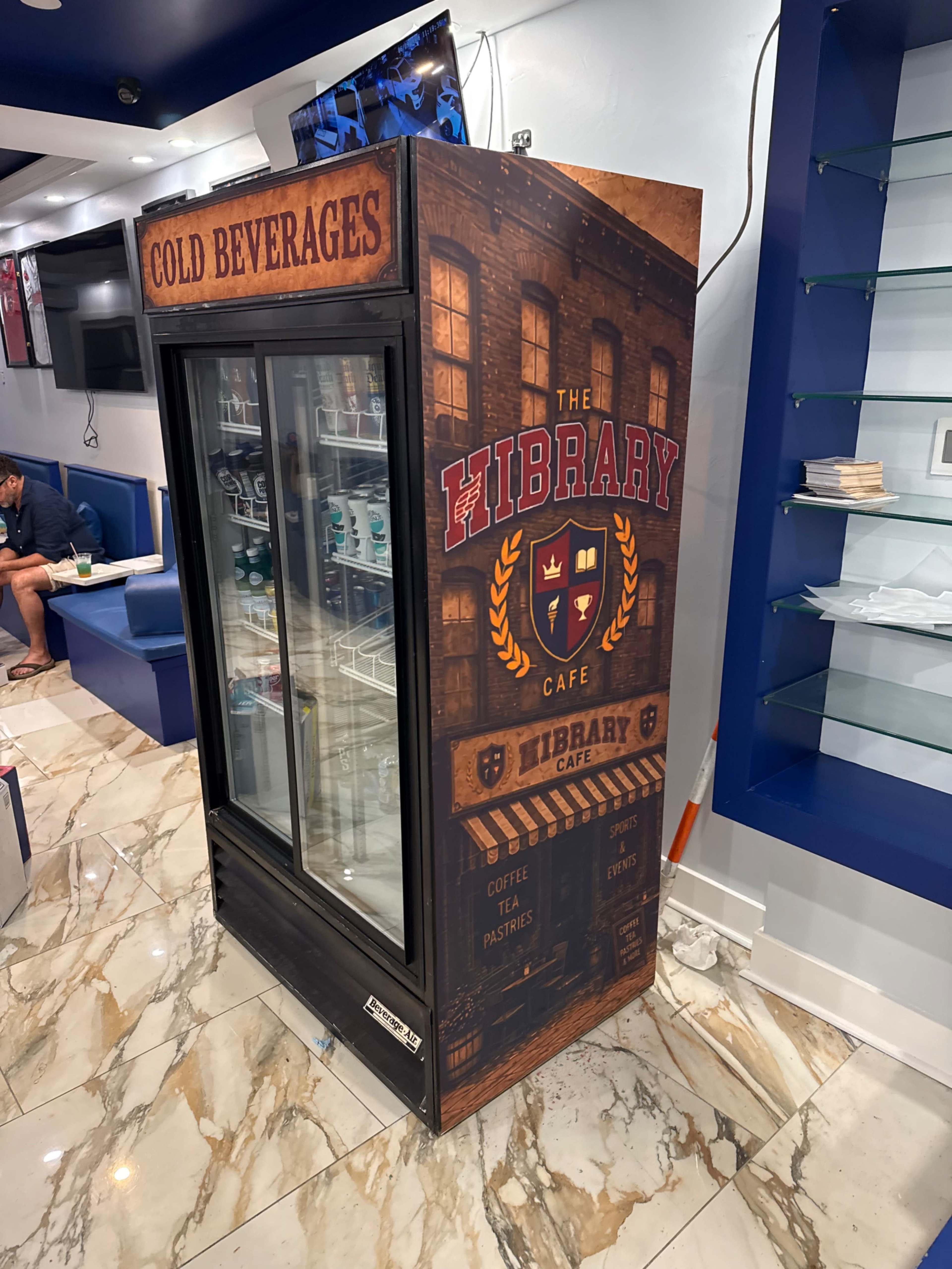 A beverage refrigerator is prominently displayed with a themed exterior featuring the words "COLD BEVERAGES" and "THE LIBRARY CAFE."