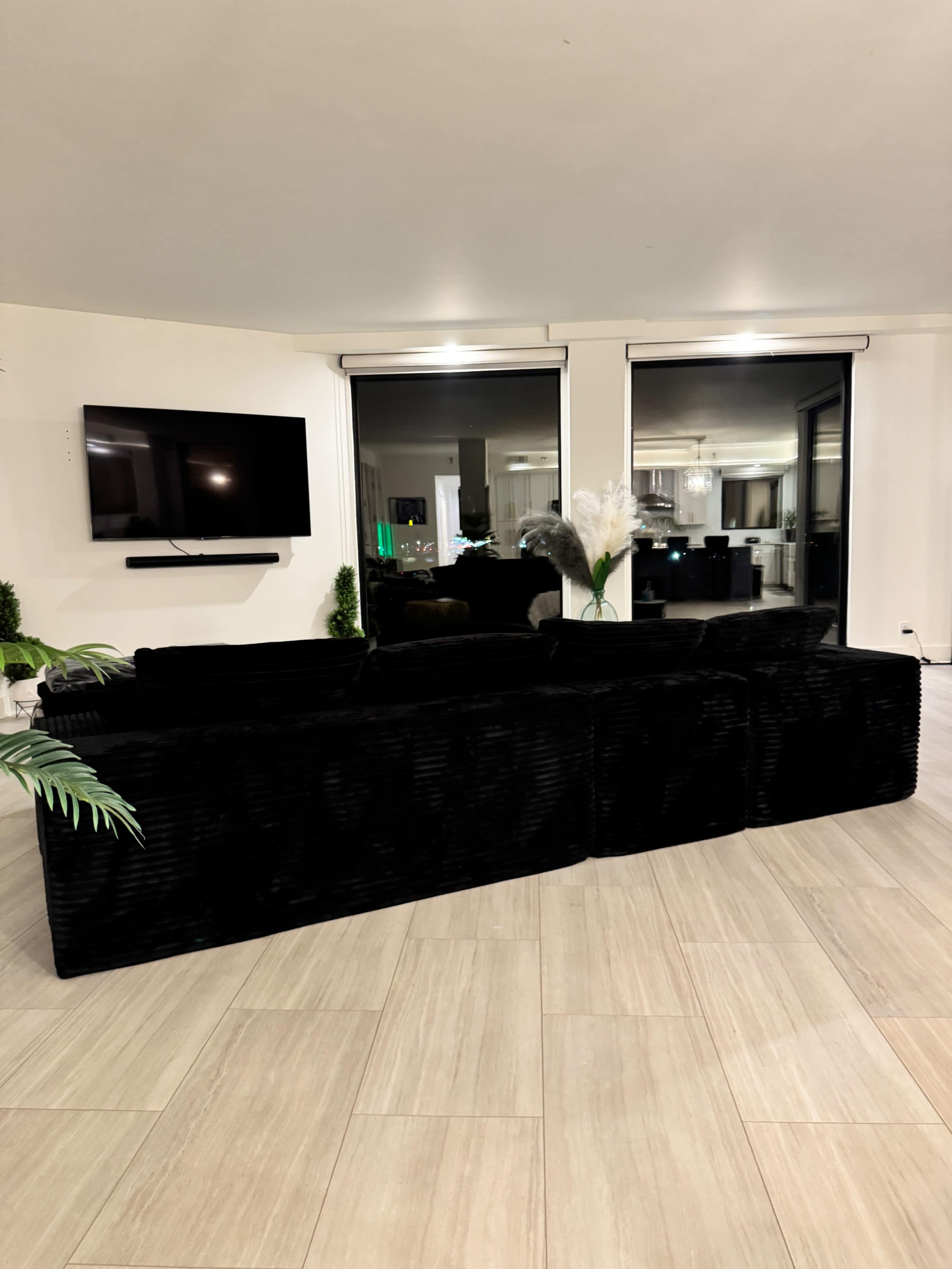 A black, plush sofa is positioned in a modern living room with large windows and a wall-mounted television.