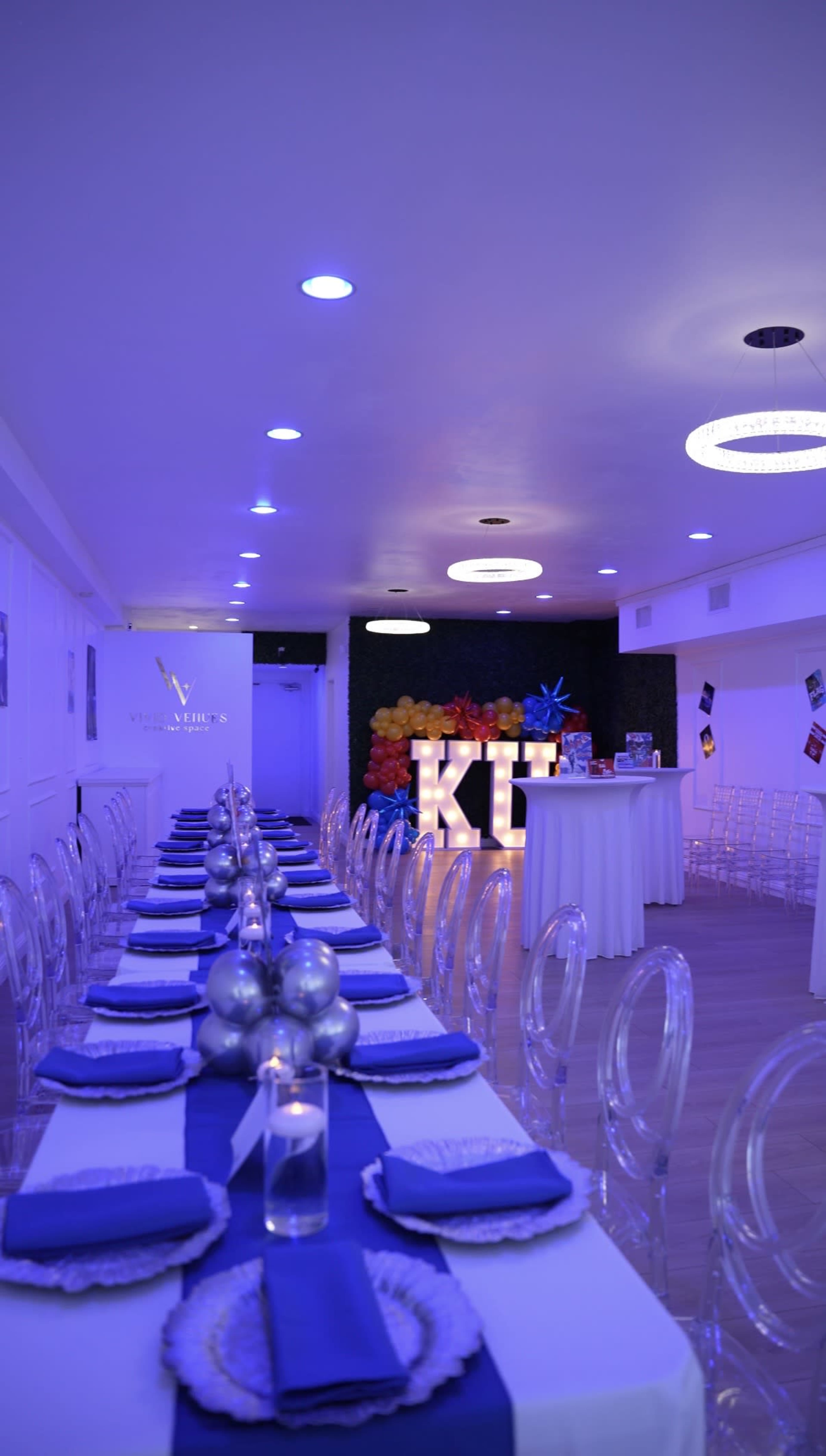 The image shows a brightly lit event space with a long table set for a gathering, decorated with blue table runners and illuminated by purple lighting.