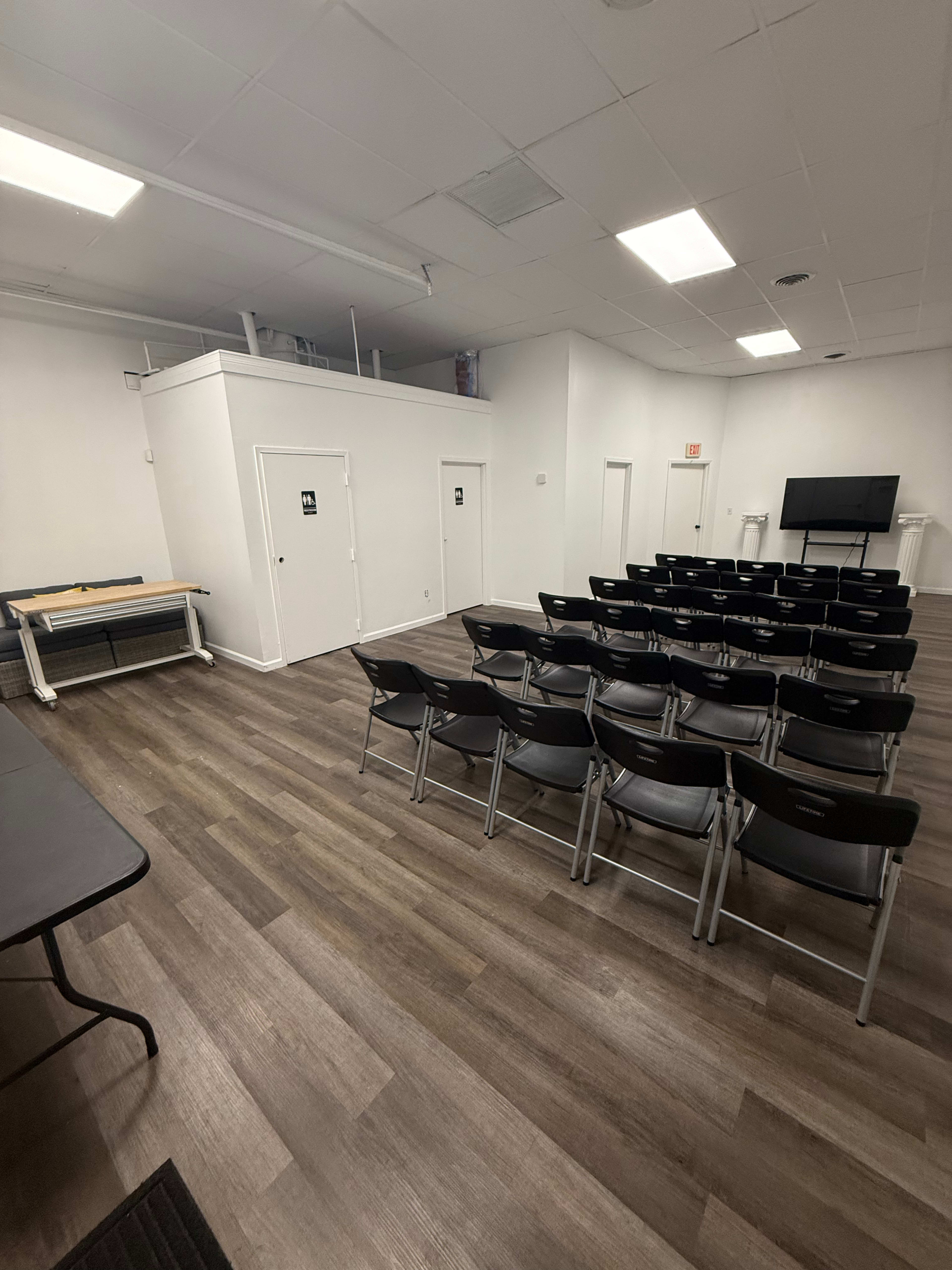 The image shows a classroom or meeting room with rows of black chairs facing a wall-mounted screen, alongside a table and two restroom doors.