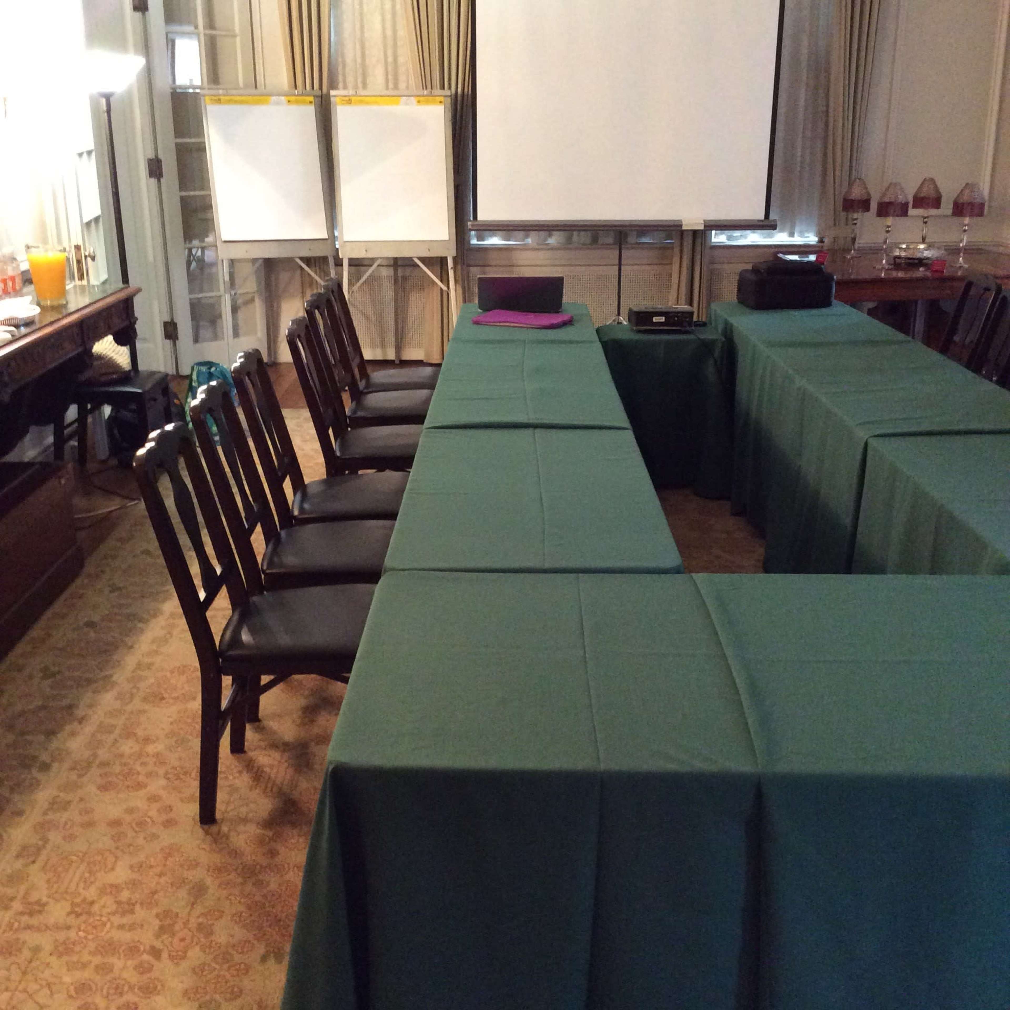 A conference room is set up with green tables arranged in a U-shape and several empty chairs facing a blank projector screen.