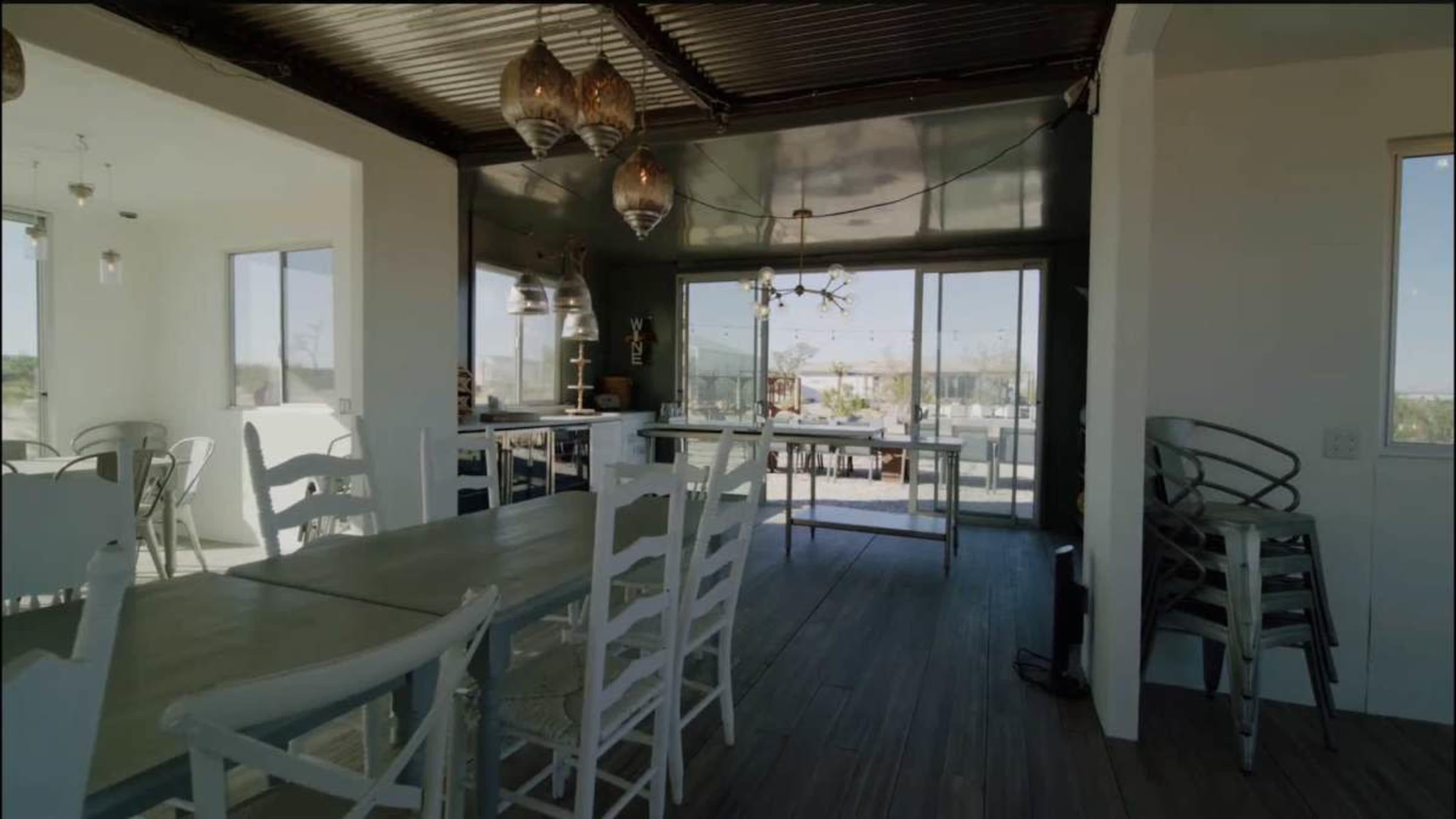 The image shows a modern dining area with white furniture, large windows, and pendant lighting, leading into an outdoor space.