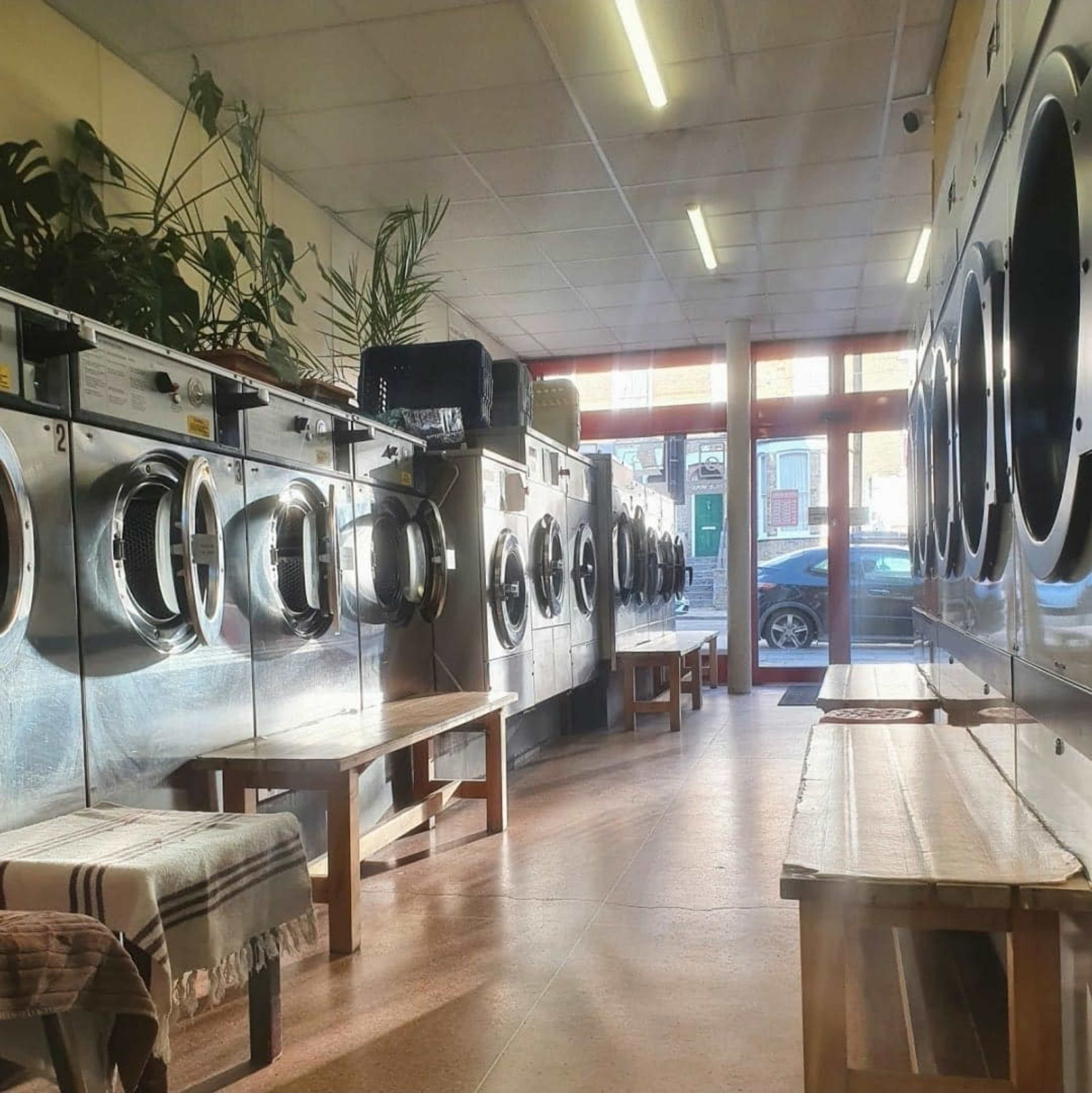 The image shows a row of washing machines lined along a laundry room with wooden benches and large windows providing natural light.