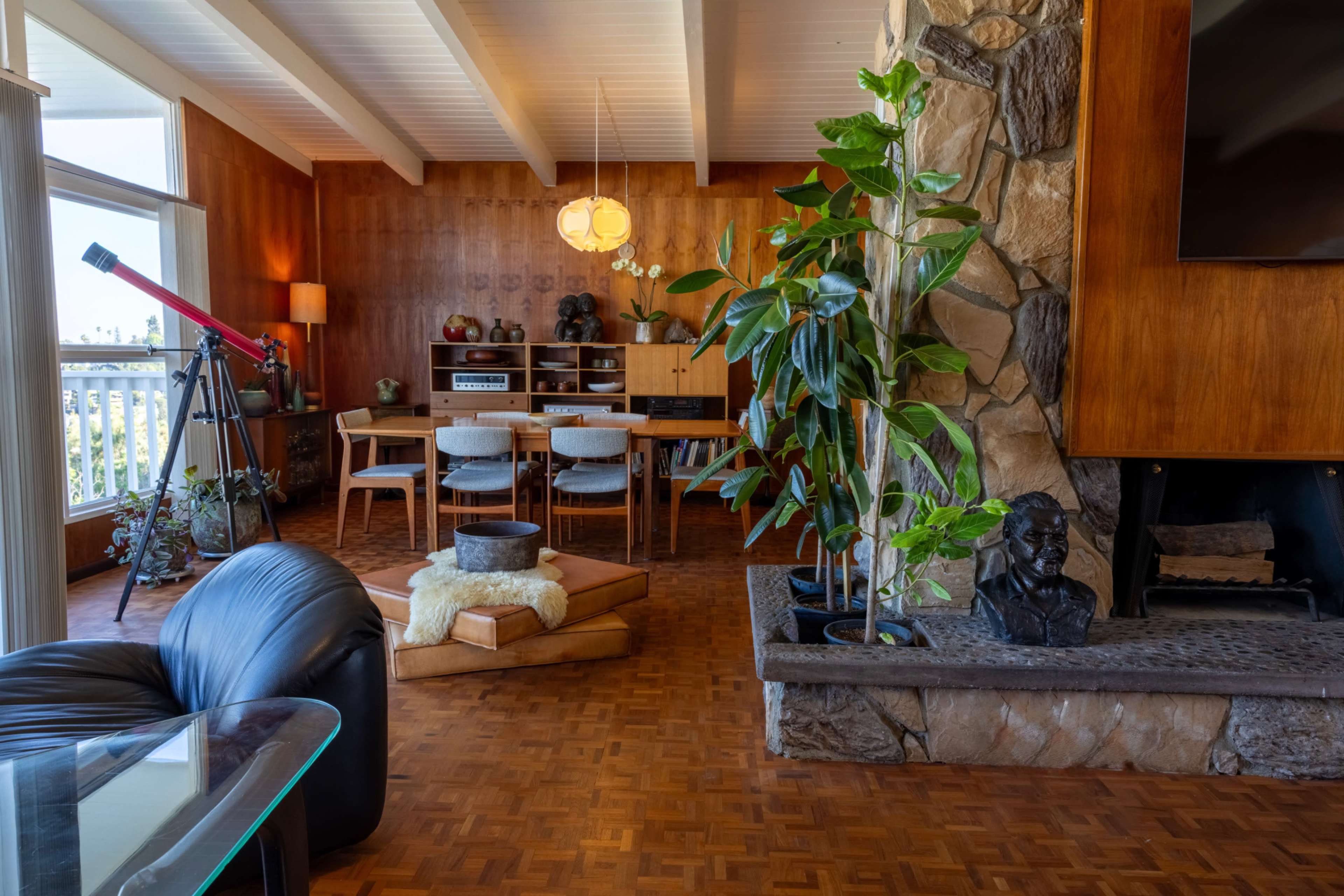 The image displays a mid-century modern living room featuring a stone fireplace, a wooden dining area with seating, a large indoor plant, and a telescope positioned by a window.