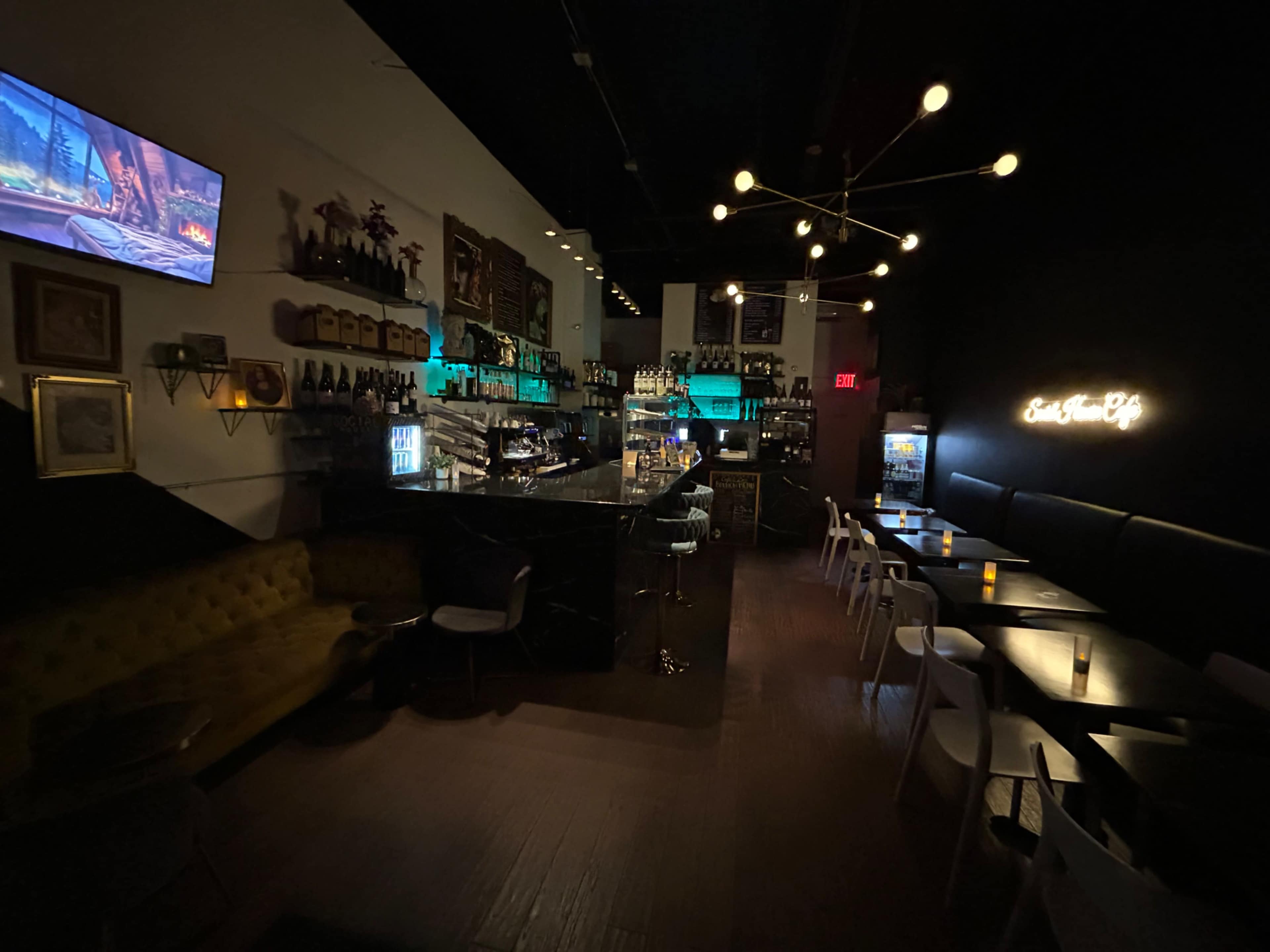 A dimly lit bar area with a counter, seating, and a decorative wall featuring bottles and a television screen.