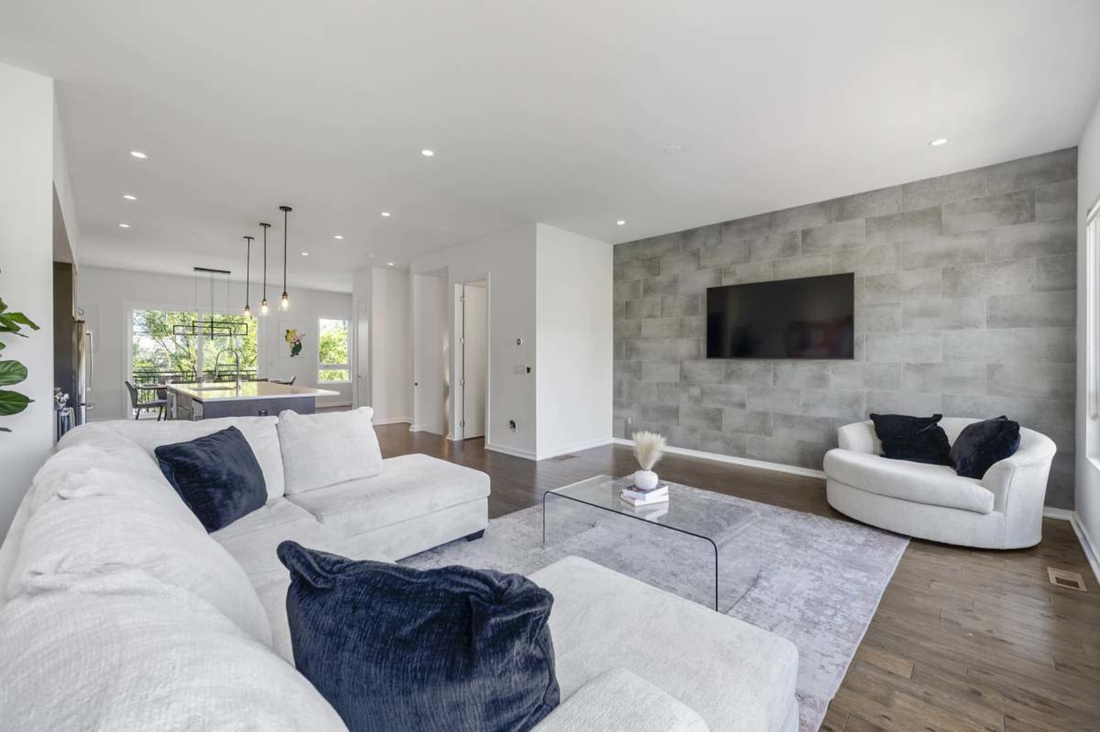 The image shows a modern living room featuring a white sectional sofa, a round chair, a glass coffee table, and a large wall-mounted television against a textured gray wall.