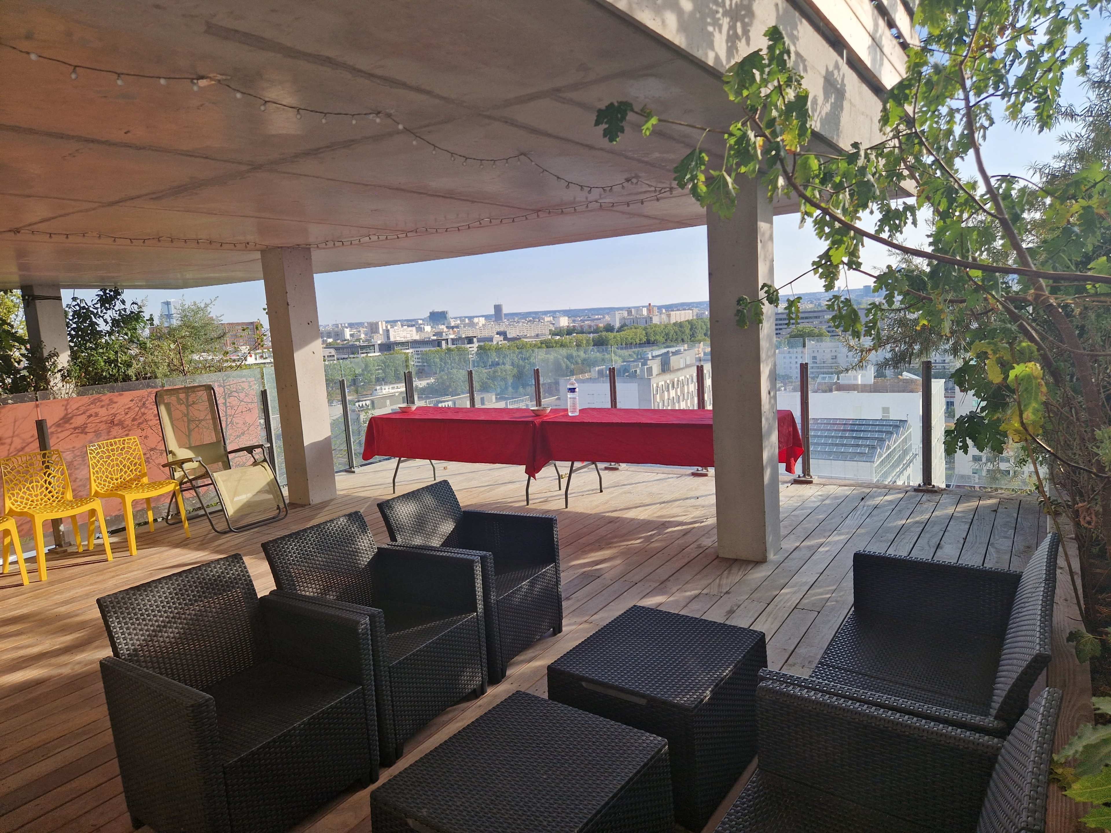 The image shows a rooftop terrace with several black wicker chairs and a long red table set against a backdrop of a city skyline.