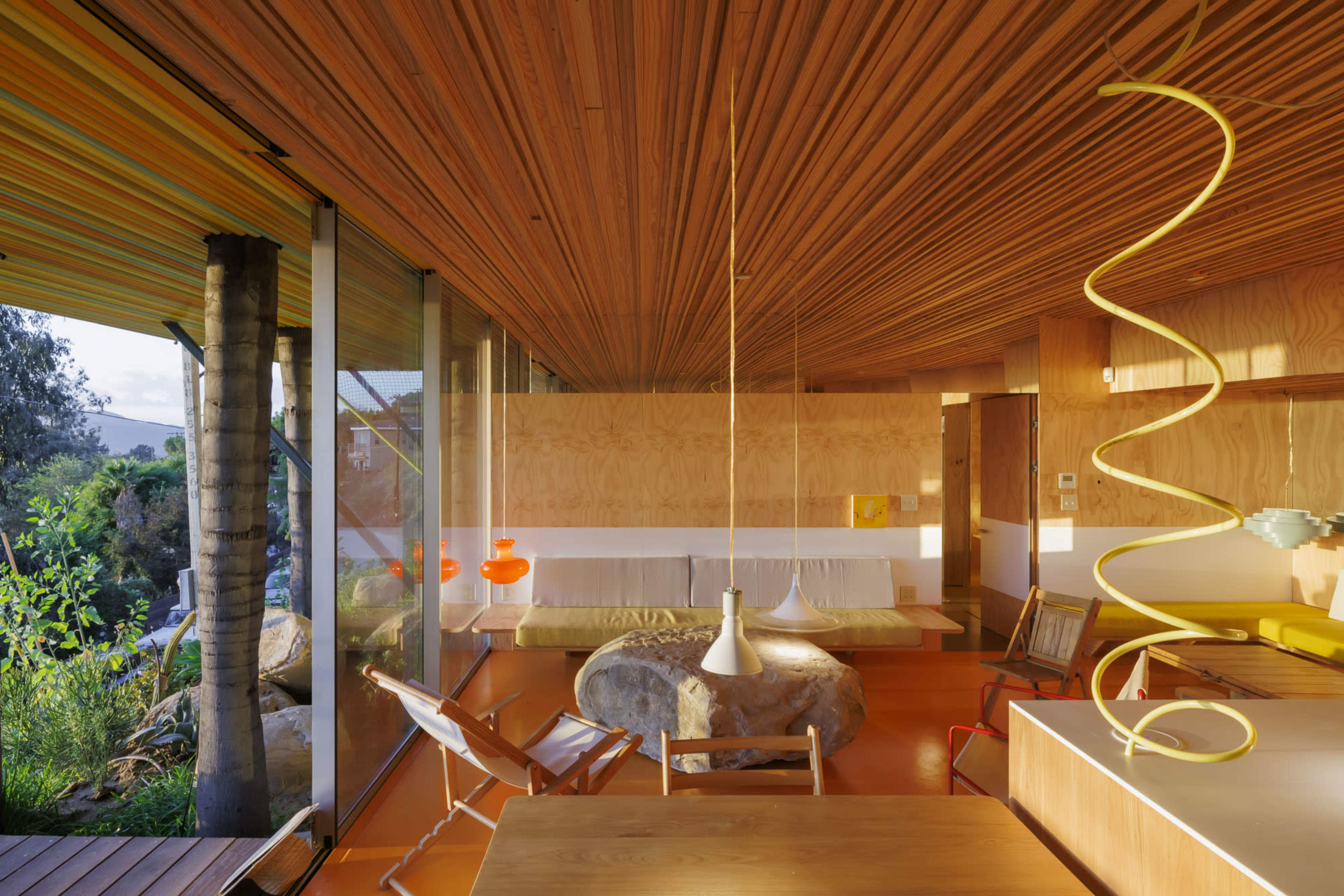 The image depicts a modern interior of a house featuring wooden ceilings, large glass windows, and a combination of contemporary furniture and natural elements.