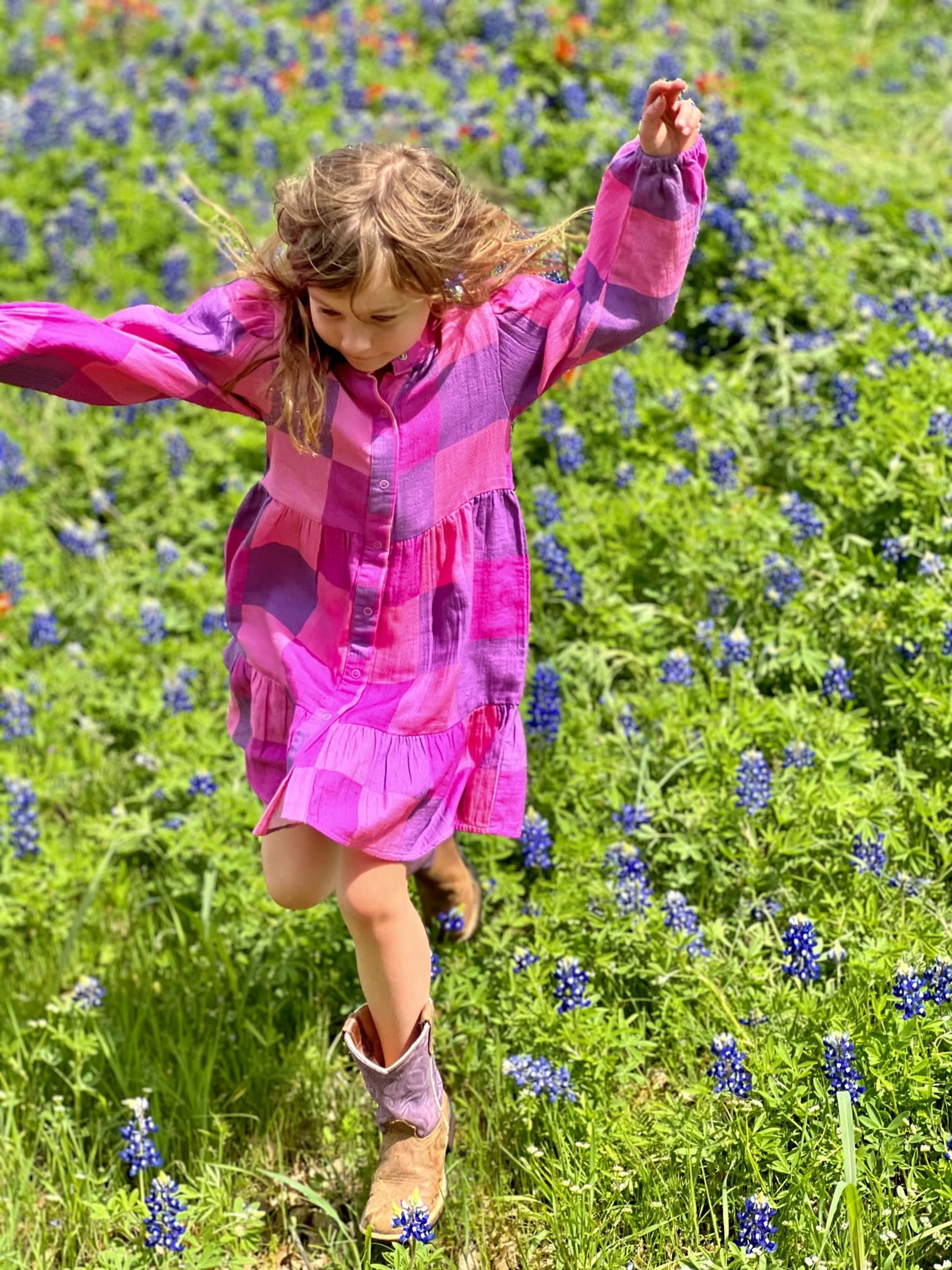 A girl in a pink checkered dress jumps through a field of bluebonnets.