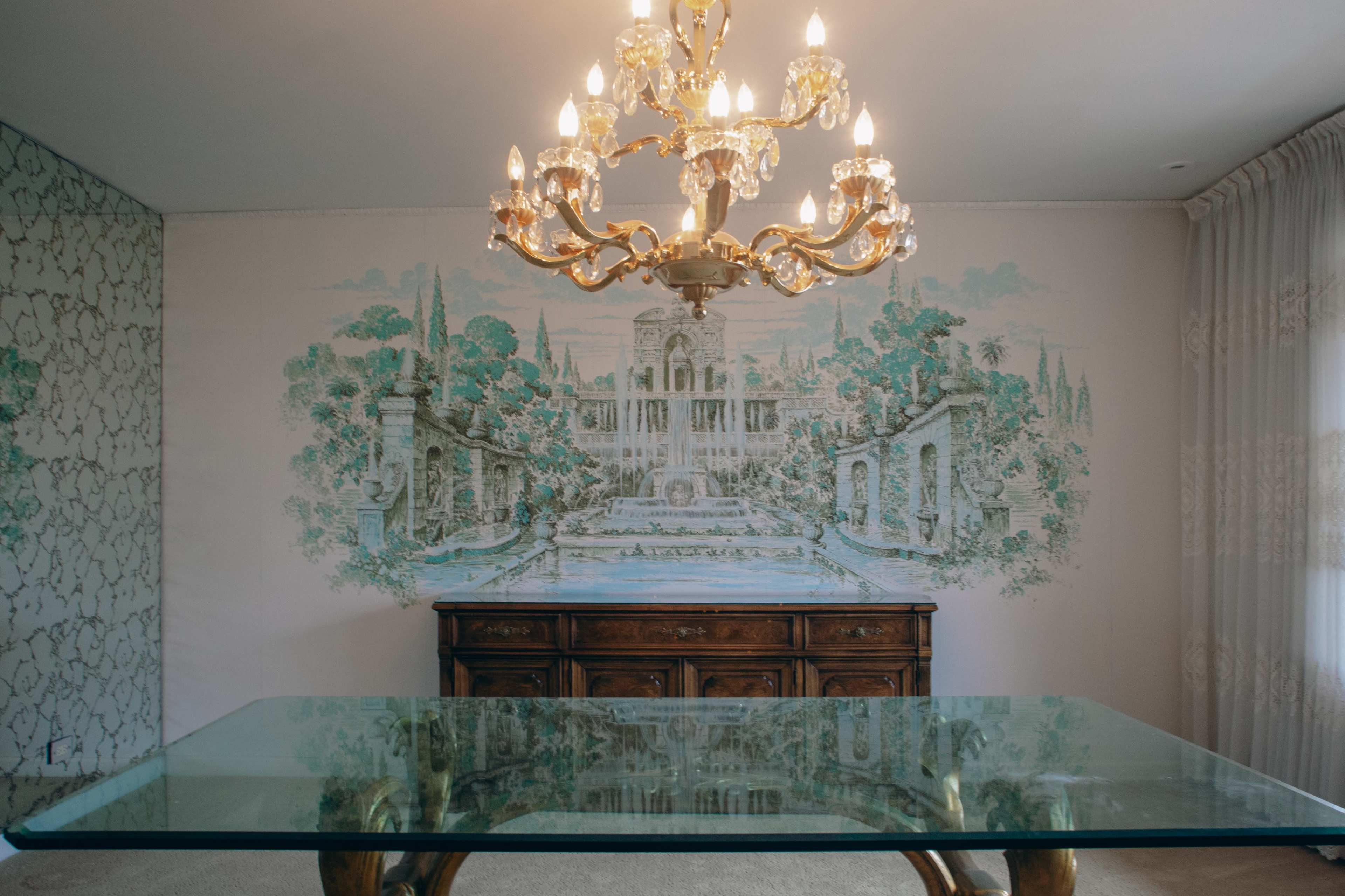The image shows a dining room with a glass table, an ornate chandelier, and a mural of a garden scene on the wall behind an antique wooden sideboard.
