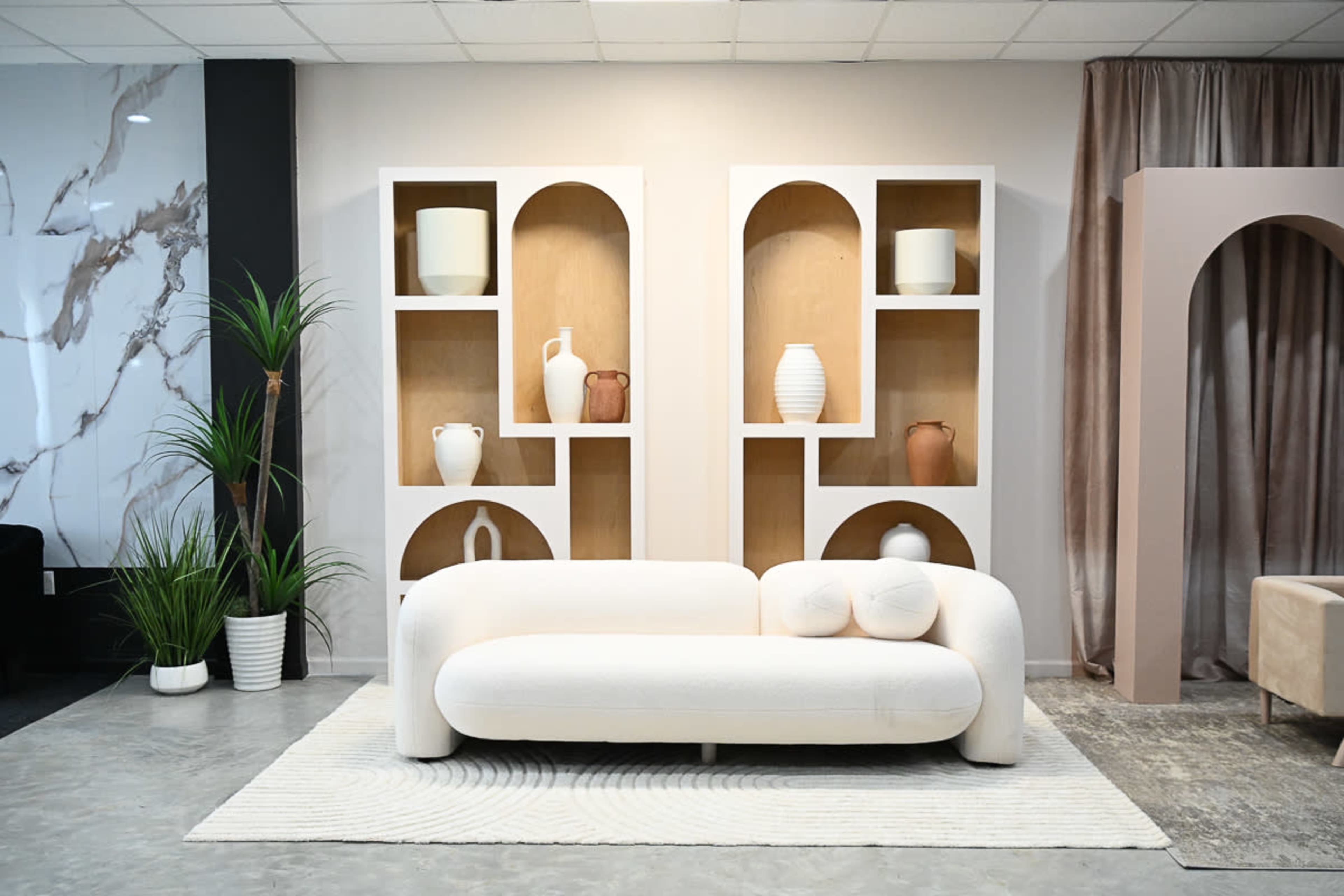 A modern living room setup featuring a white curved sofa in front of two shelving units with decorative items and plants.