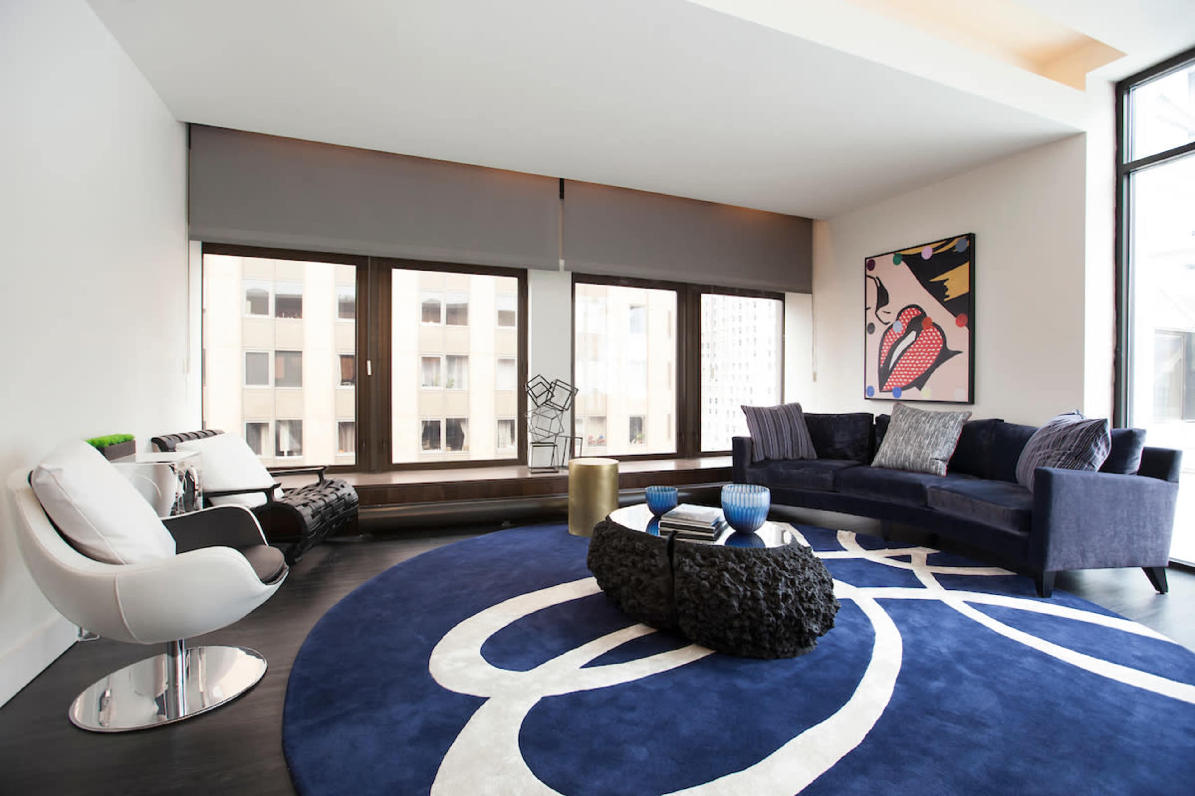 A modern living room with large windows, a round blue area rug, a dark sofa, white armchairs, and an abstract artwork on the wall.