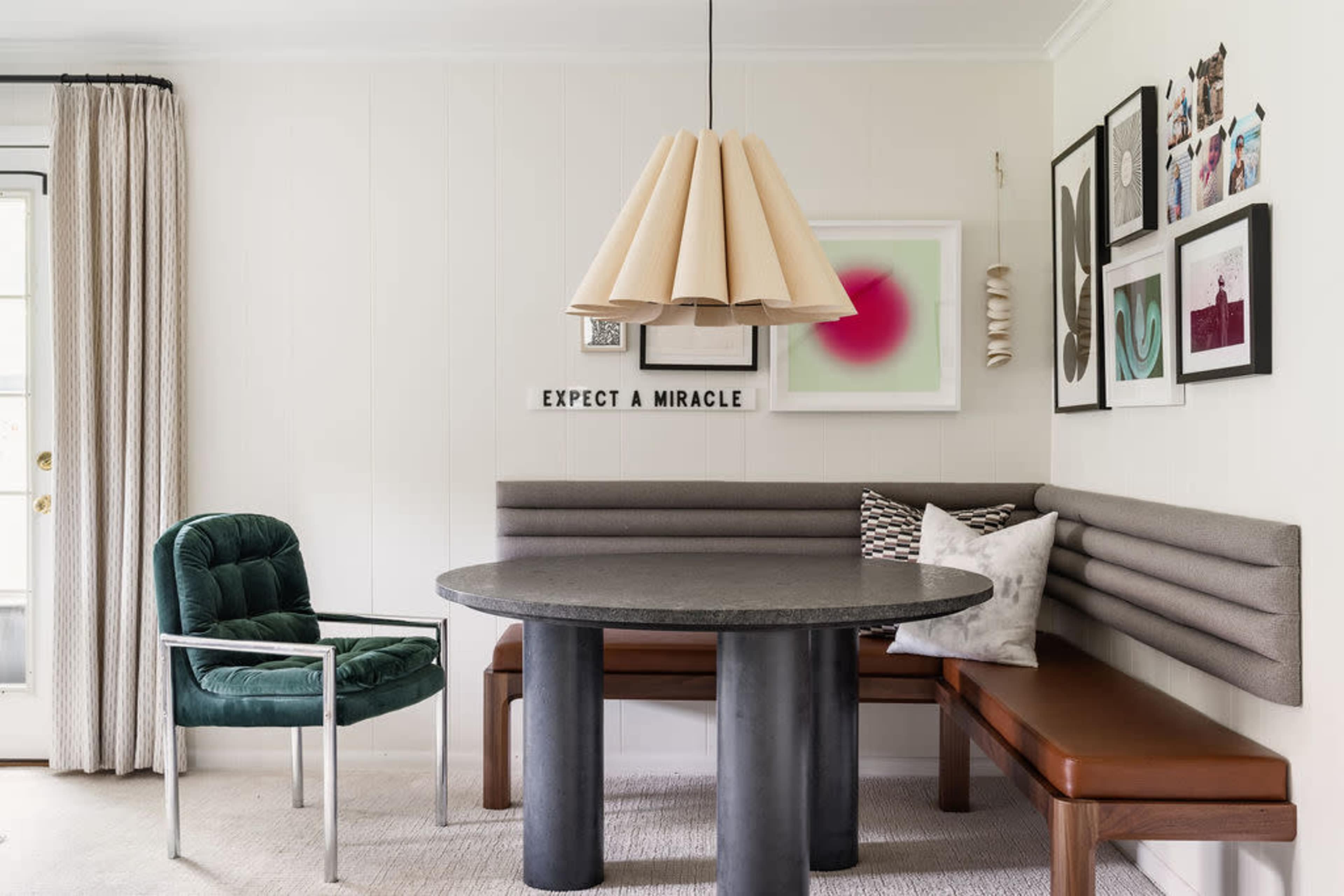 A modern dining area with a round table, a bench with backrest seating, and a green chair, adorned with several framed artworks on the wall.