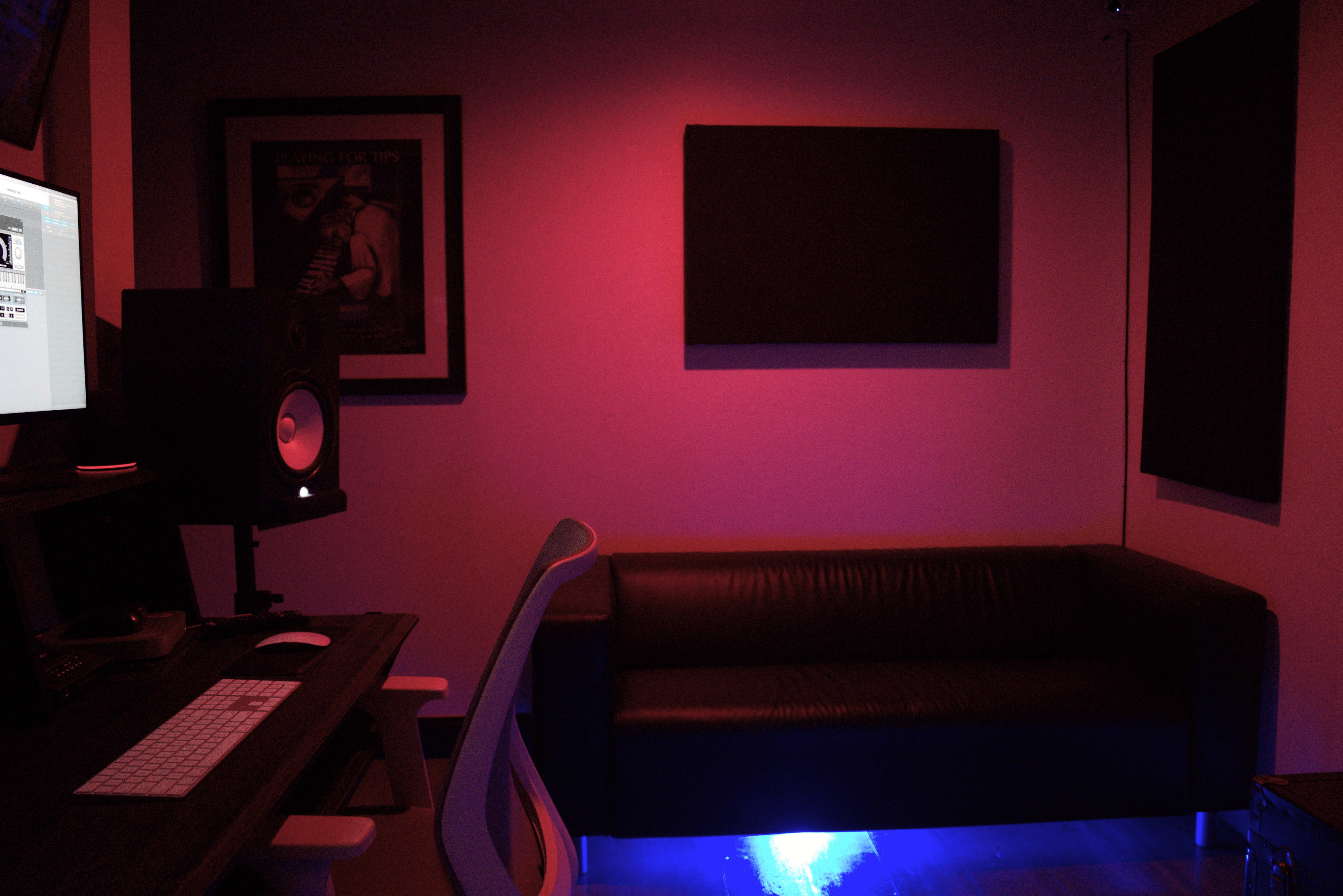 A music studio features a darkened space with a computer desk, an audio speaker, and a black couch illuminated by blue lighting.