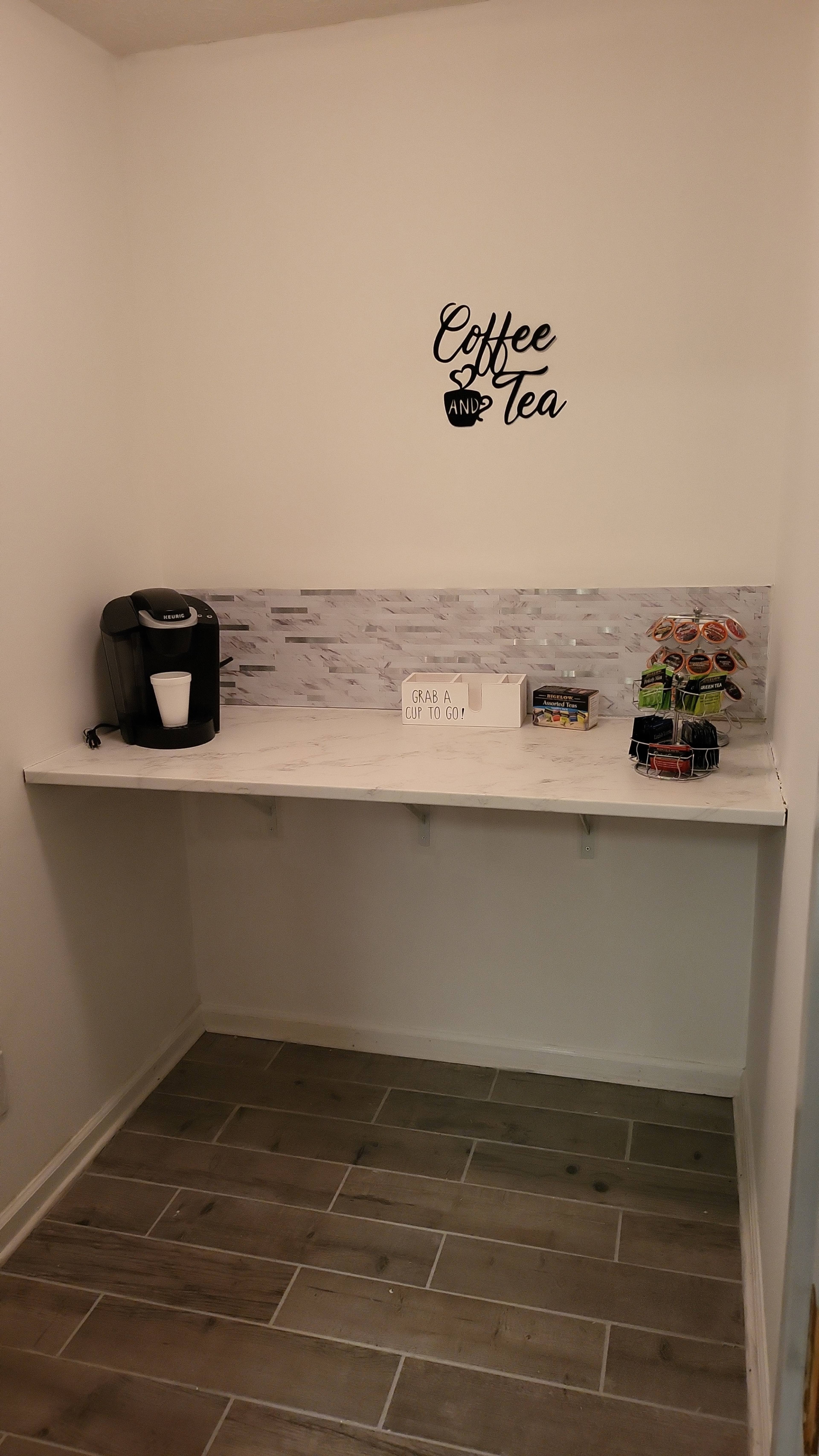 A small nook features a coffee maker and a selection of snacks against a white wall with the words "Coffee and Tea" above a marble-inspired countertop.
