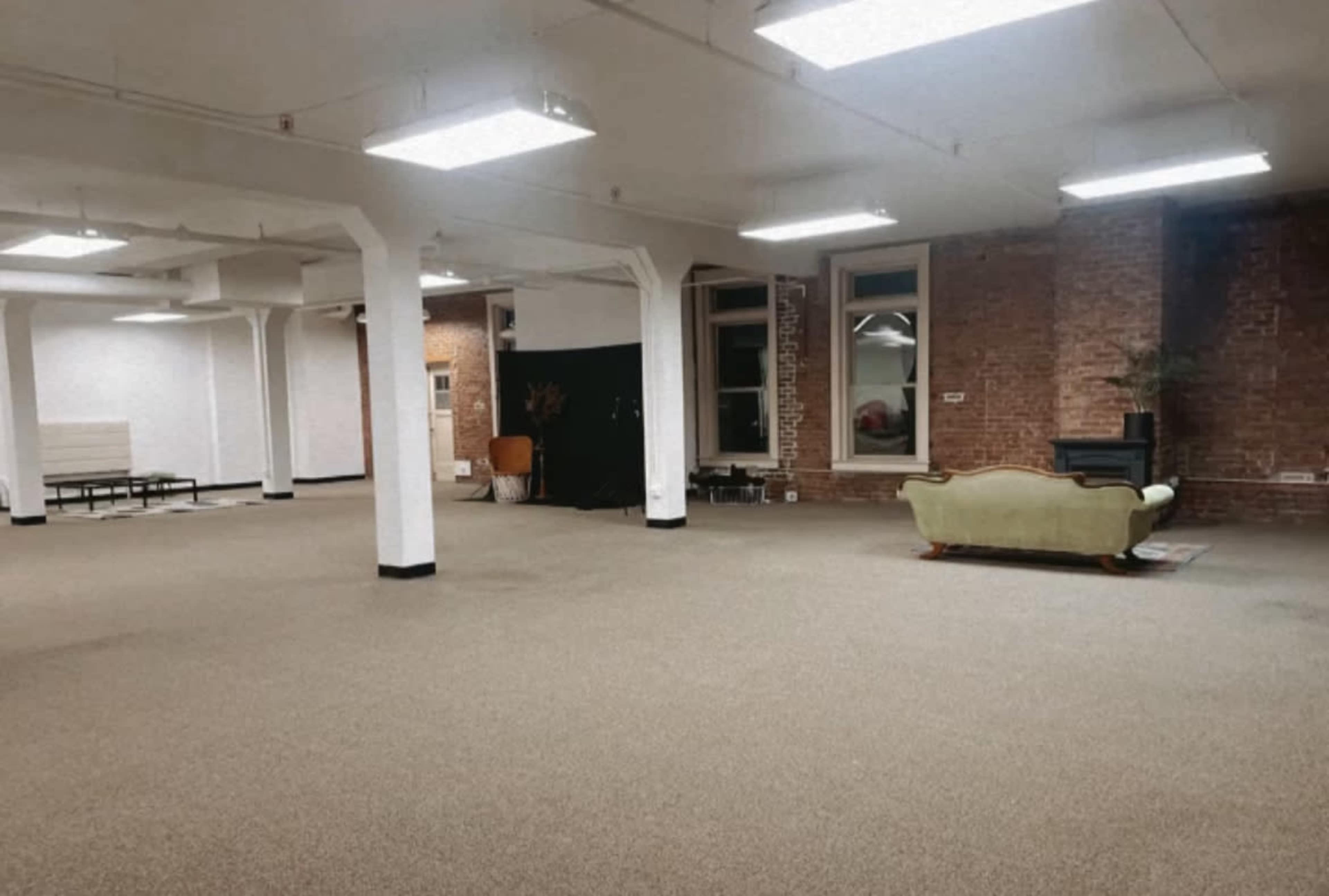 An empty room with carpeted flooring, brick walls, and overhead fluorescent lights contains a vintage green sofa and a few scattered furniture pieces.