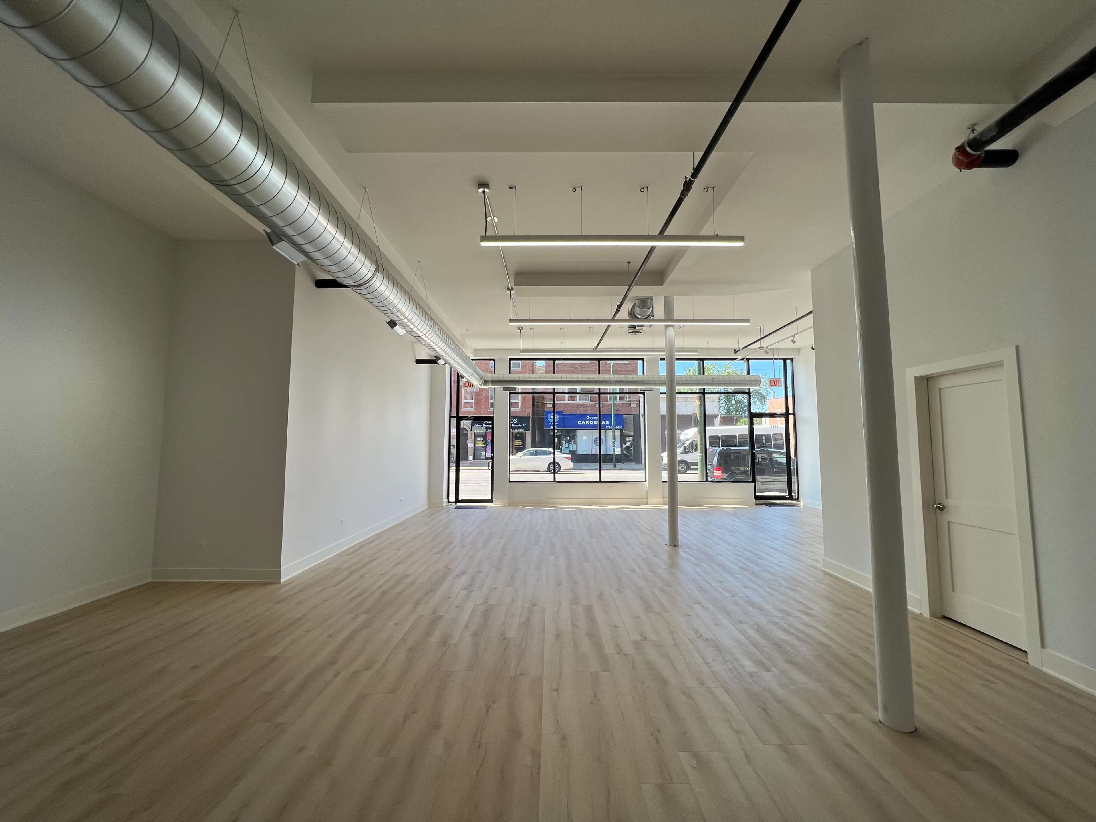 A spacious, empty interior of a retail space with large windows, light wooden flooring, and exposed ductwork on the ceiling.