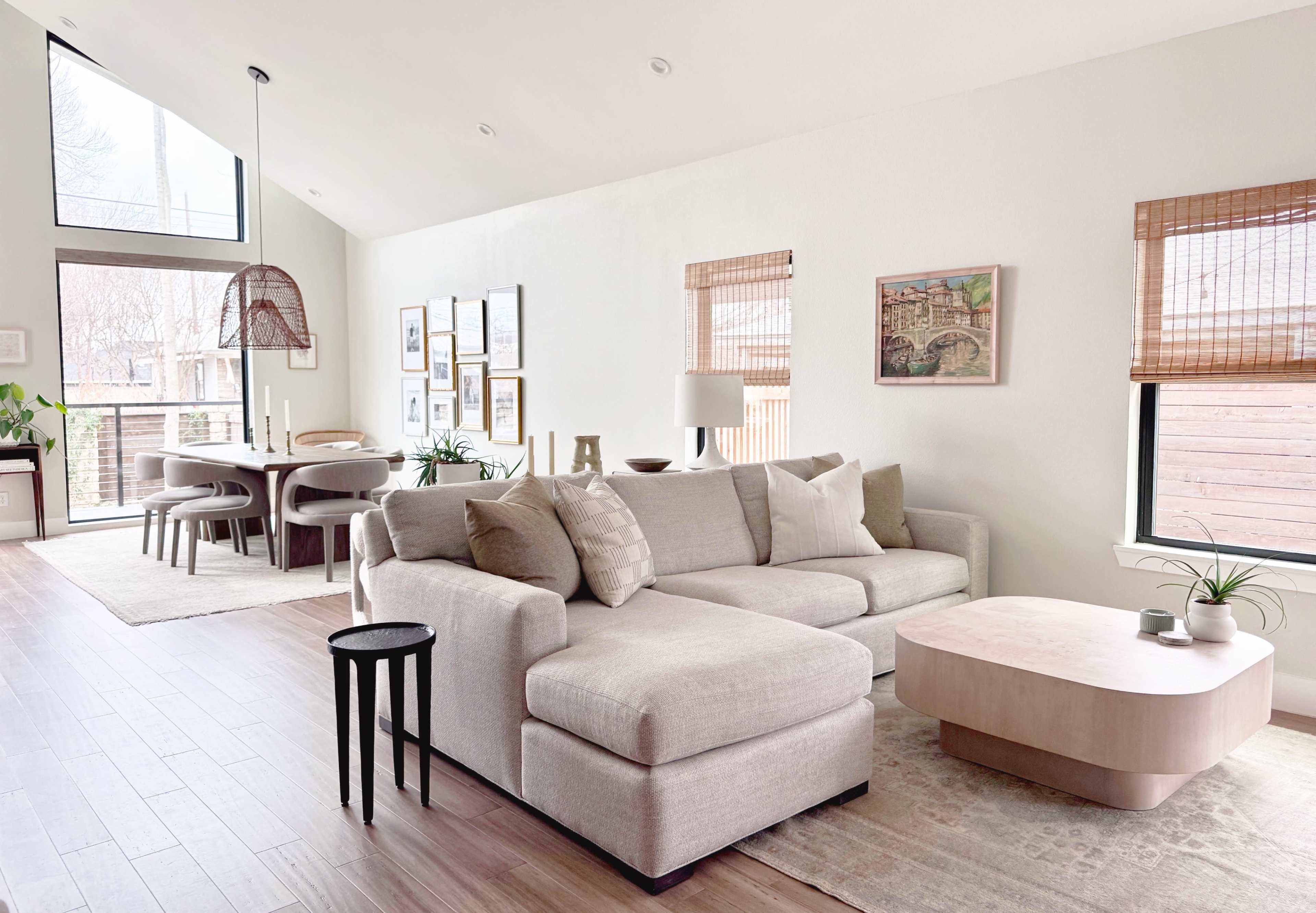 The image shows a spacious living room with a light-colored sectional sofa, a wooden coffee table, and a dining area featuring a round table and several framed artworks on the walls.