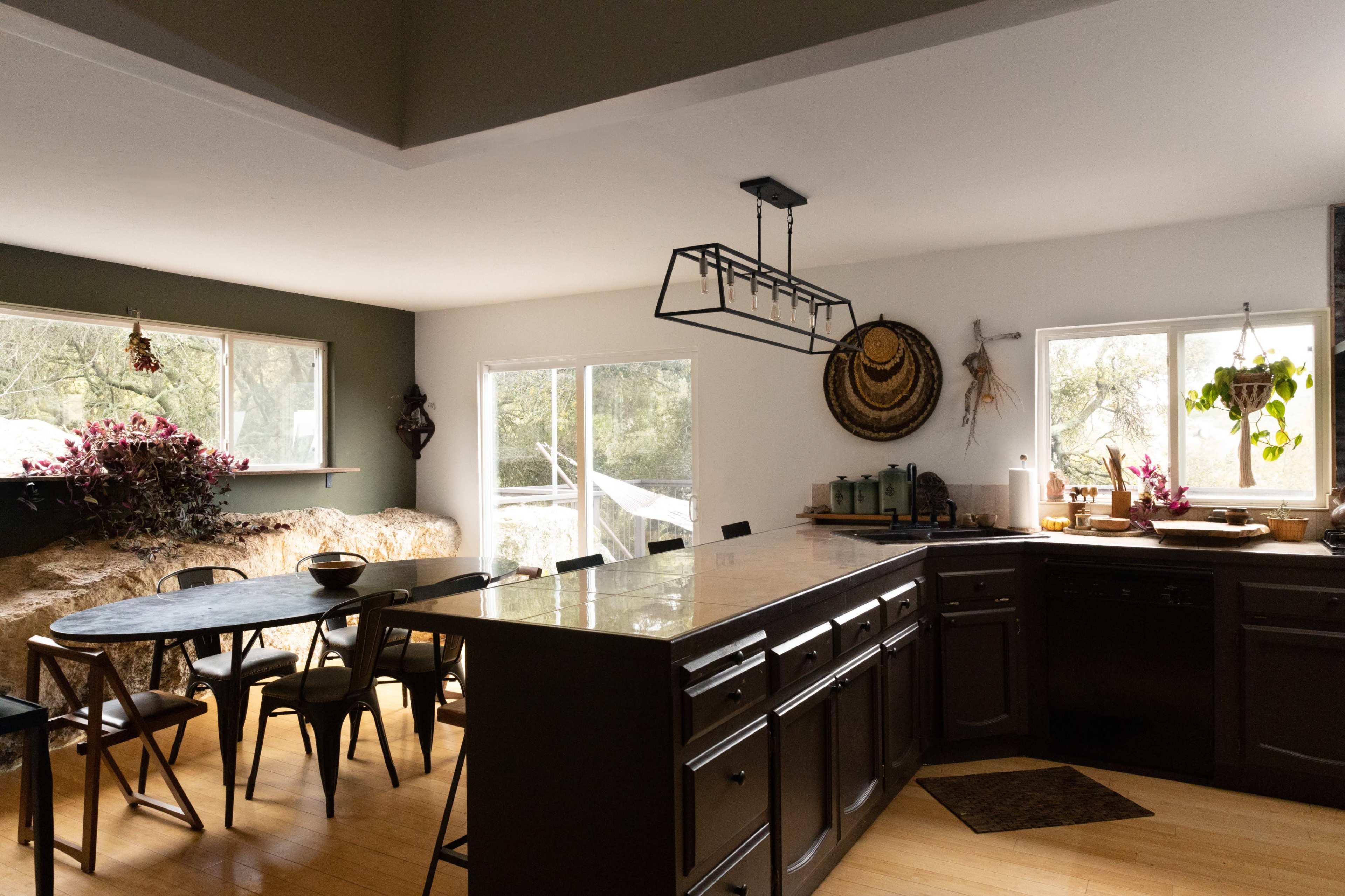 The image shows a modern kitchen with a large island, black cabinetry, a dining area with a table and chairs, and large windows allowing natural light.