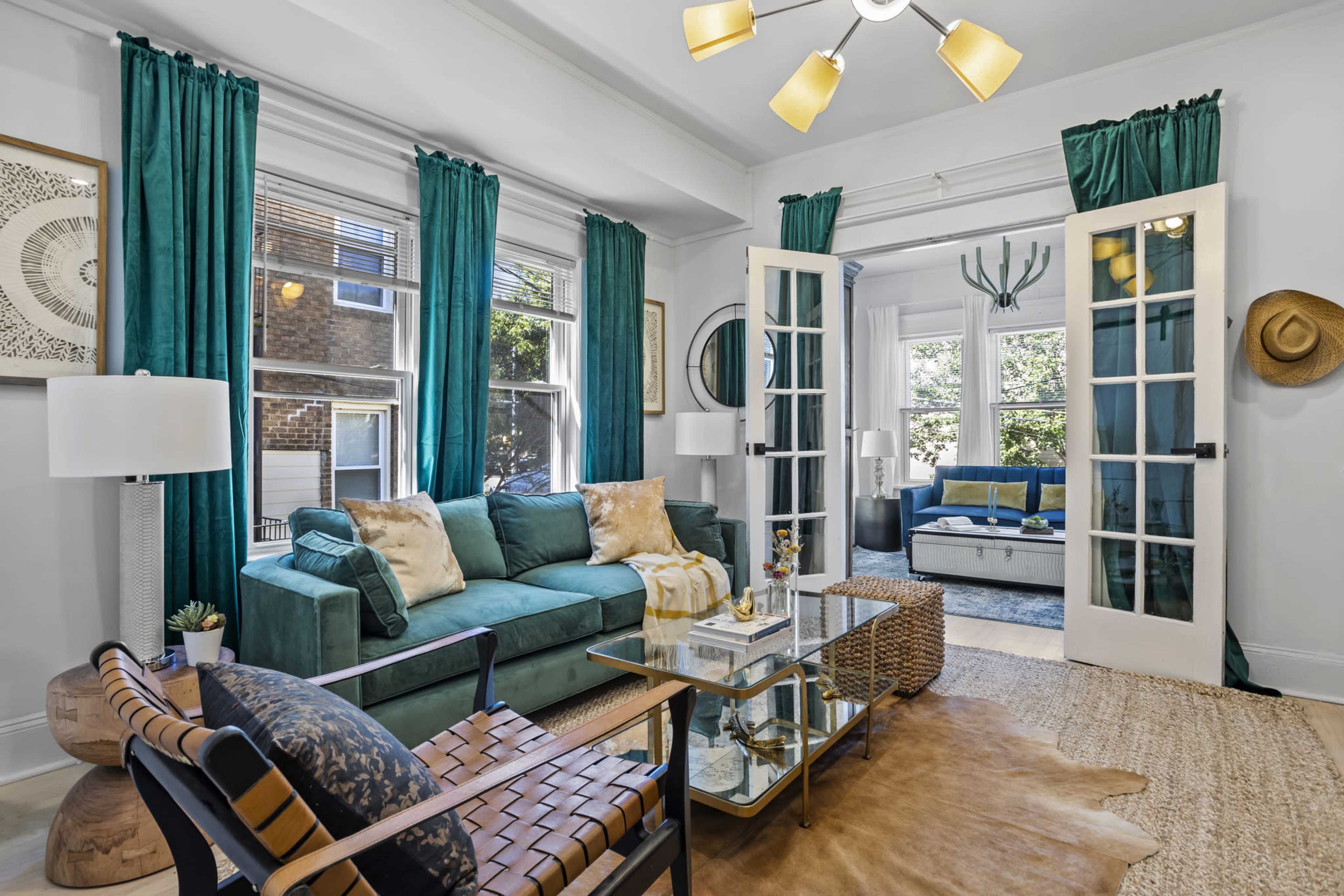A bright living room featuring a teal sofa, a glass coffee table, and large windows with green curtains.