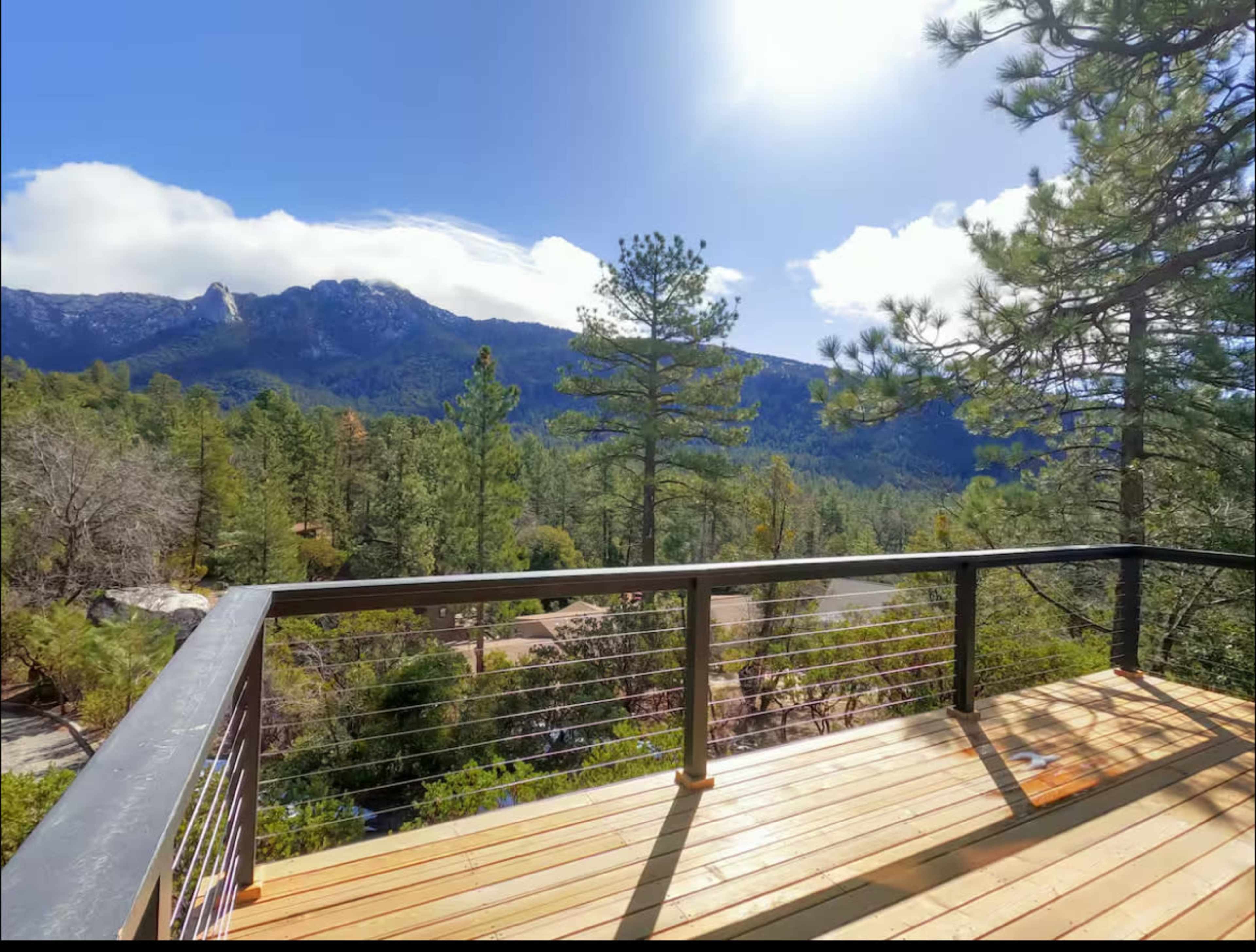 A wooden deck overlooks a forested landscape and mountains under a bright blue sky.