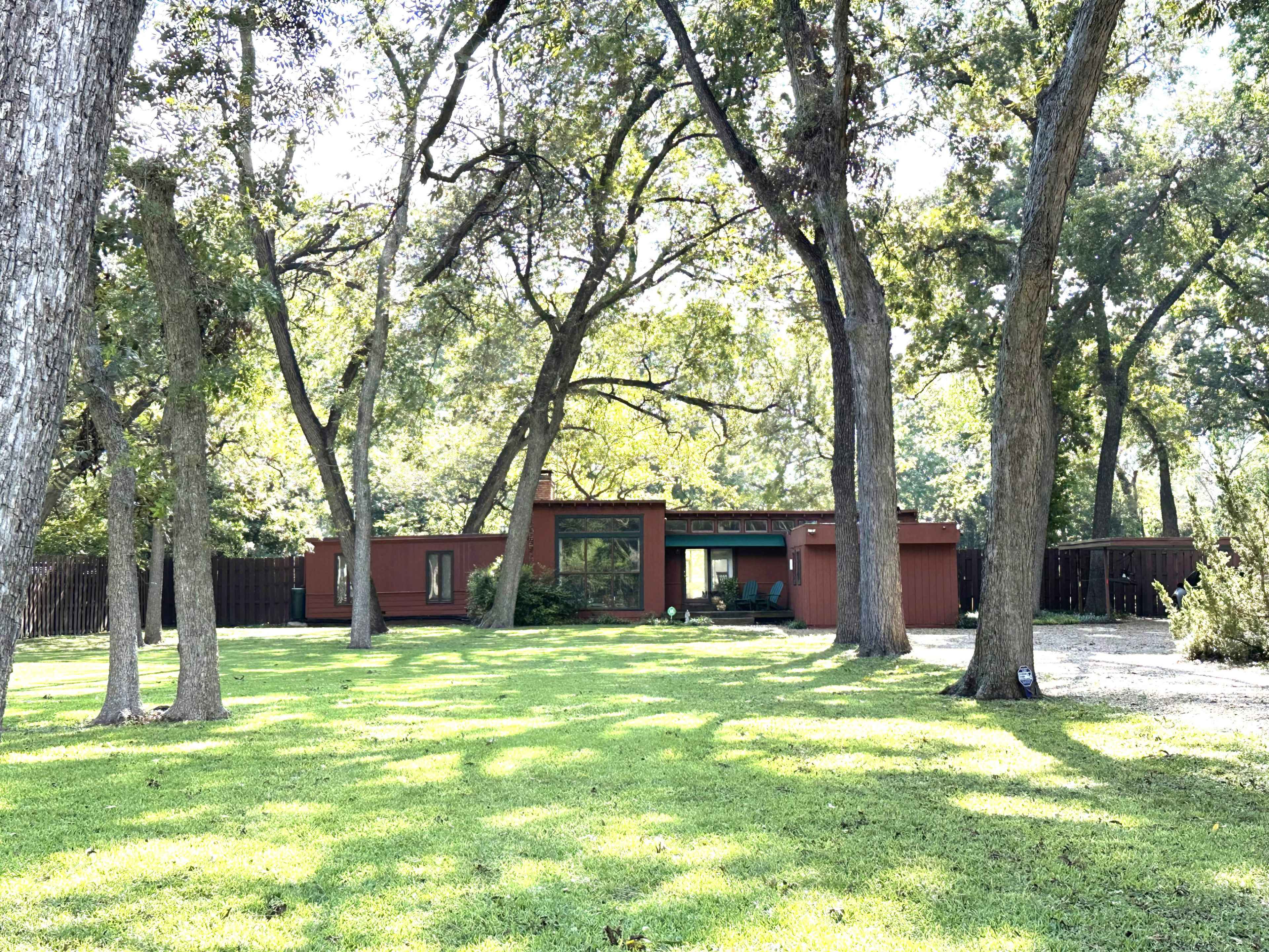 A modern flat-roofed house with large windows is situated among several tall trees on a grassy lawn.