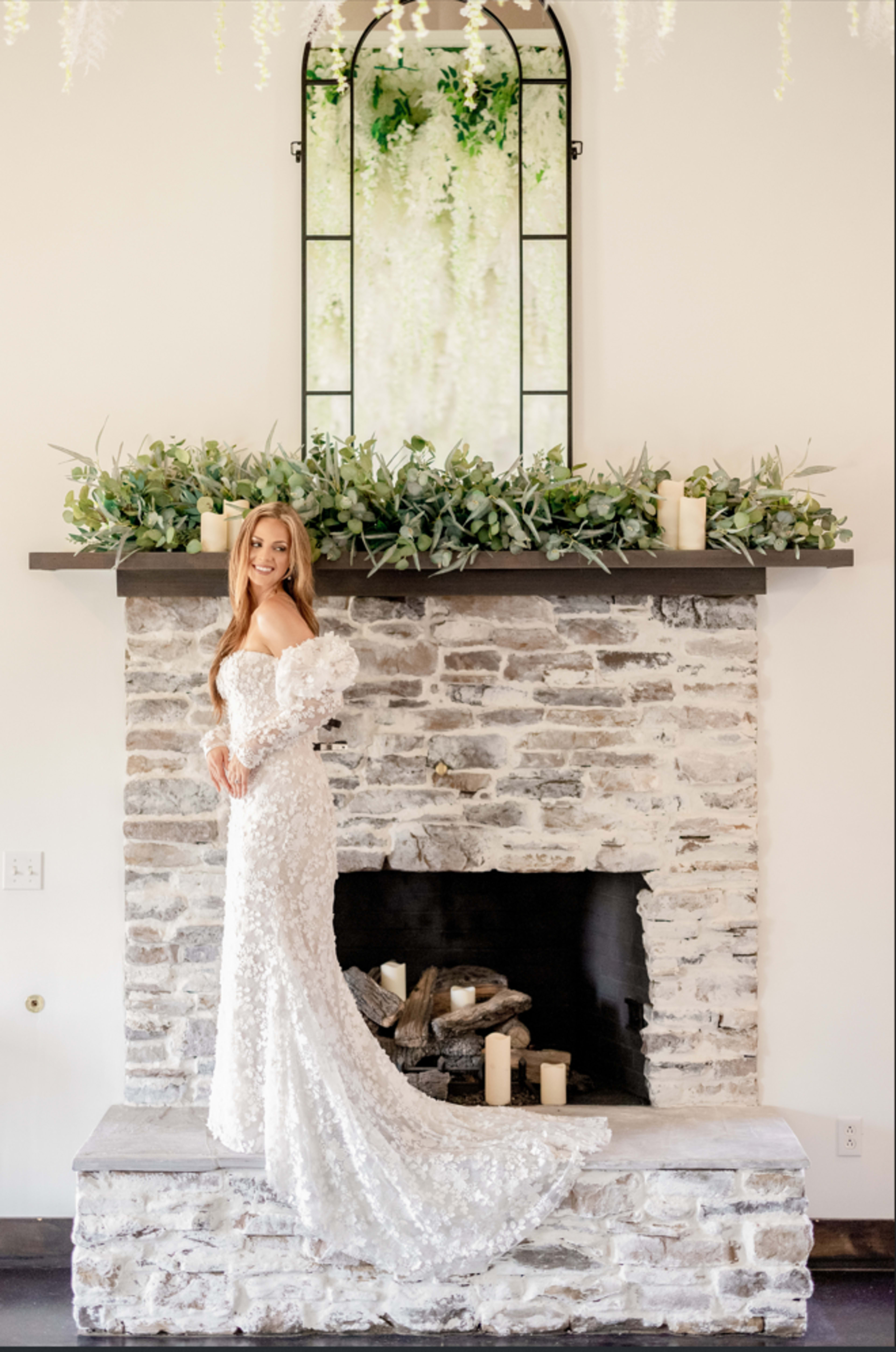 A bride in a white gown stands gracefully on a stone fireplace, with greenery and candles arranged decoratively along the mantel.