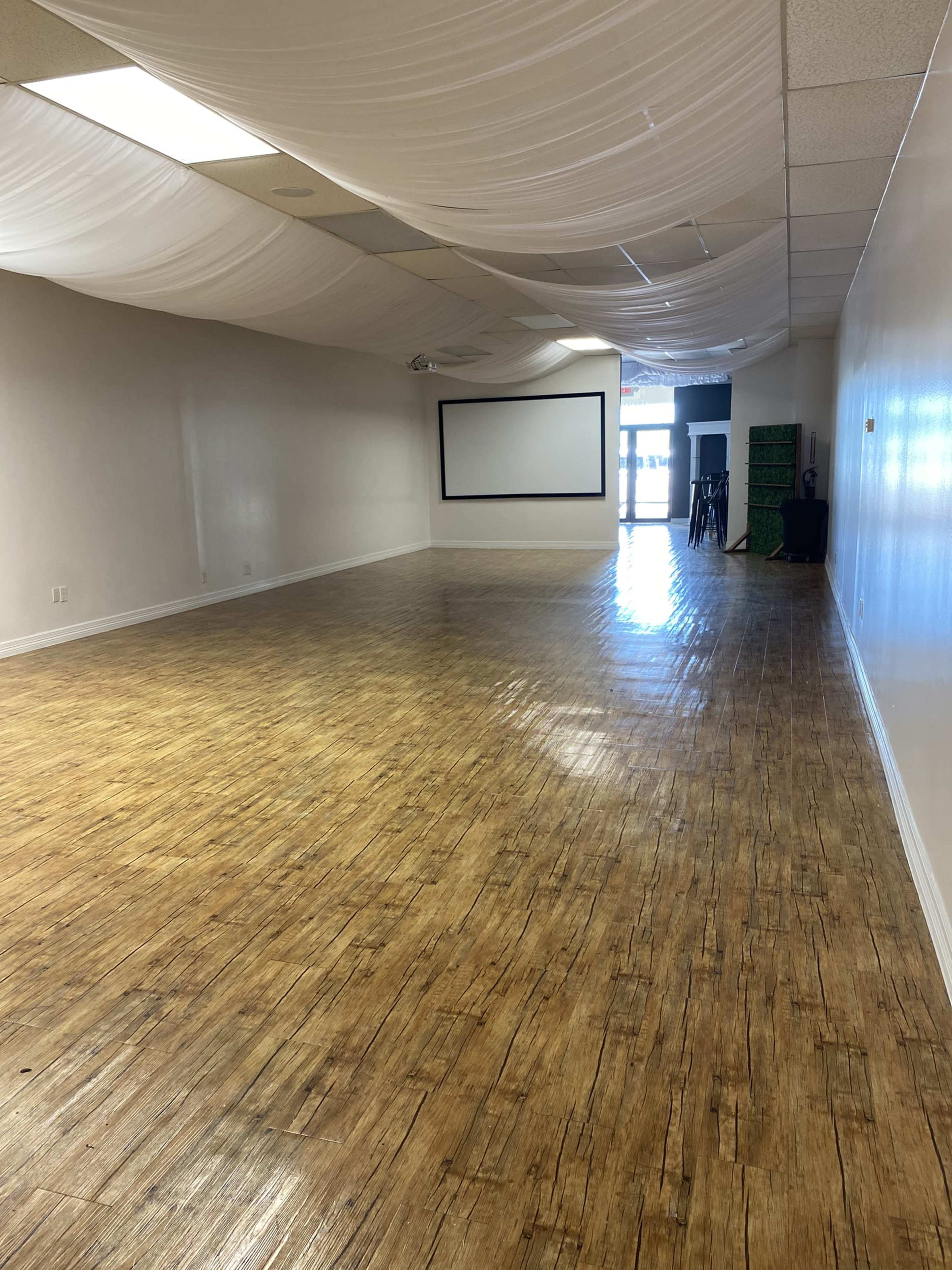 The image shows a spacious, empty room with wooden flooring, a large screen on one wall, and draped ceiling fabric.