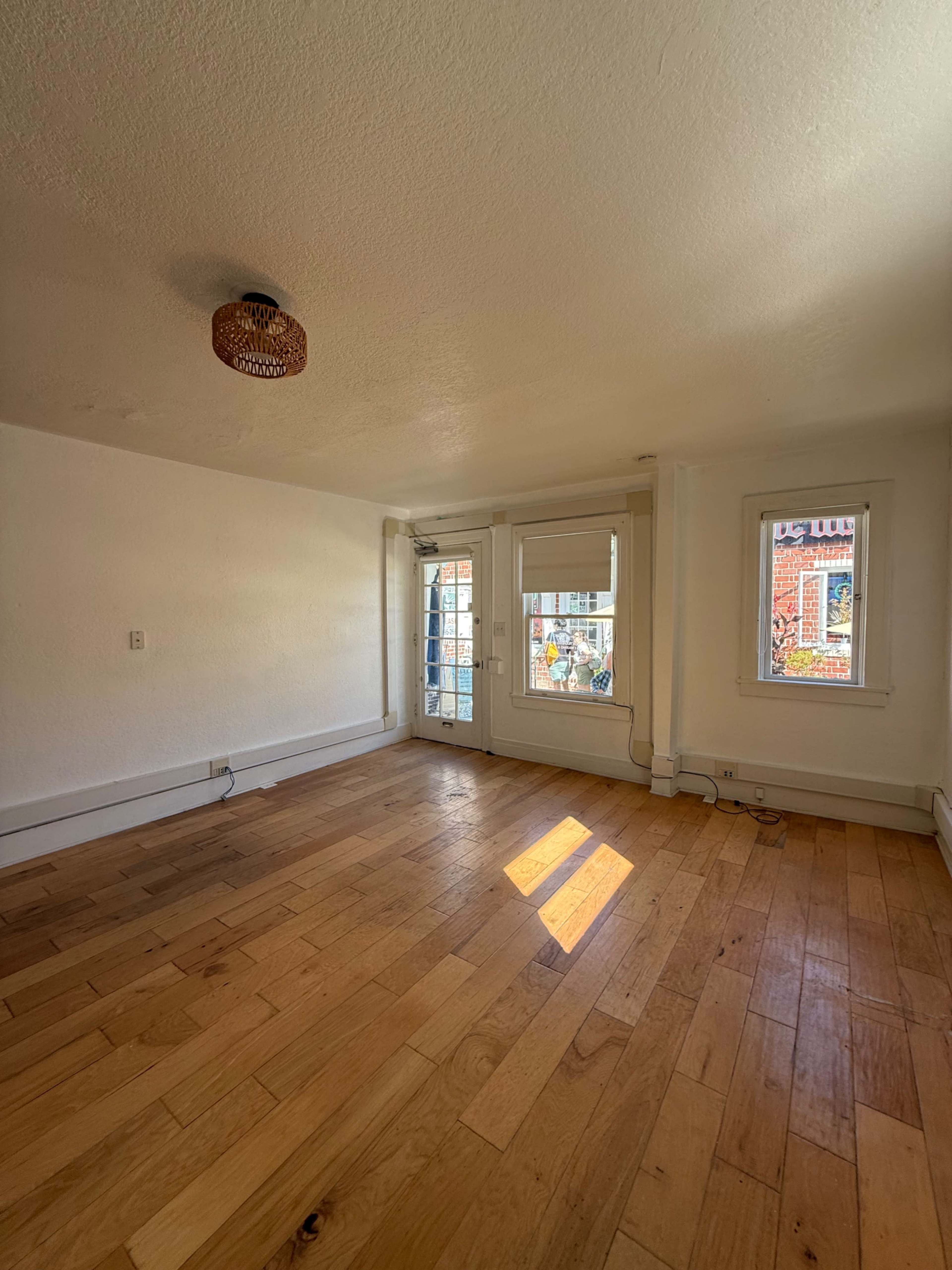 The image shows a vacant room with wooden flooring, a ceiling light fixture, and two windows letting in natural light.