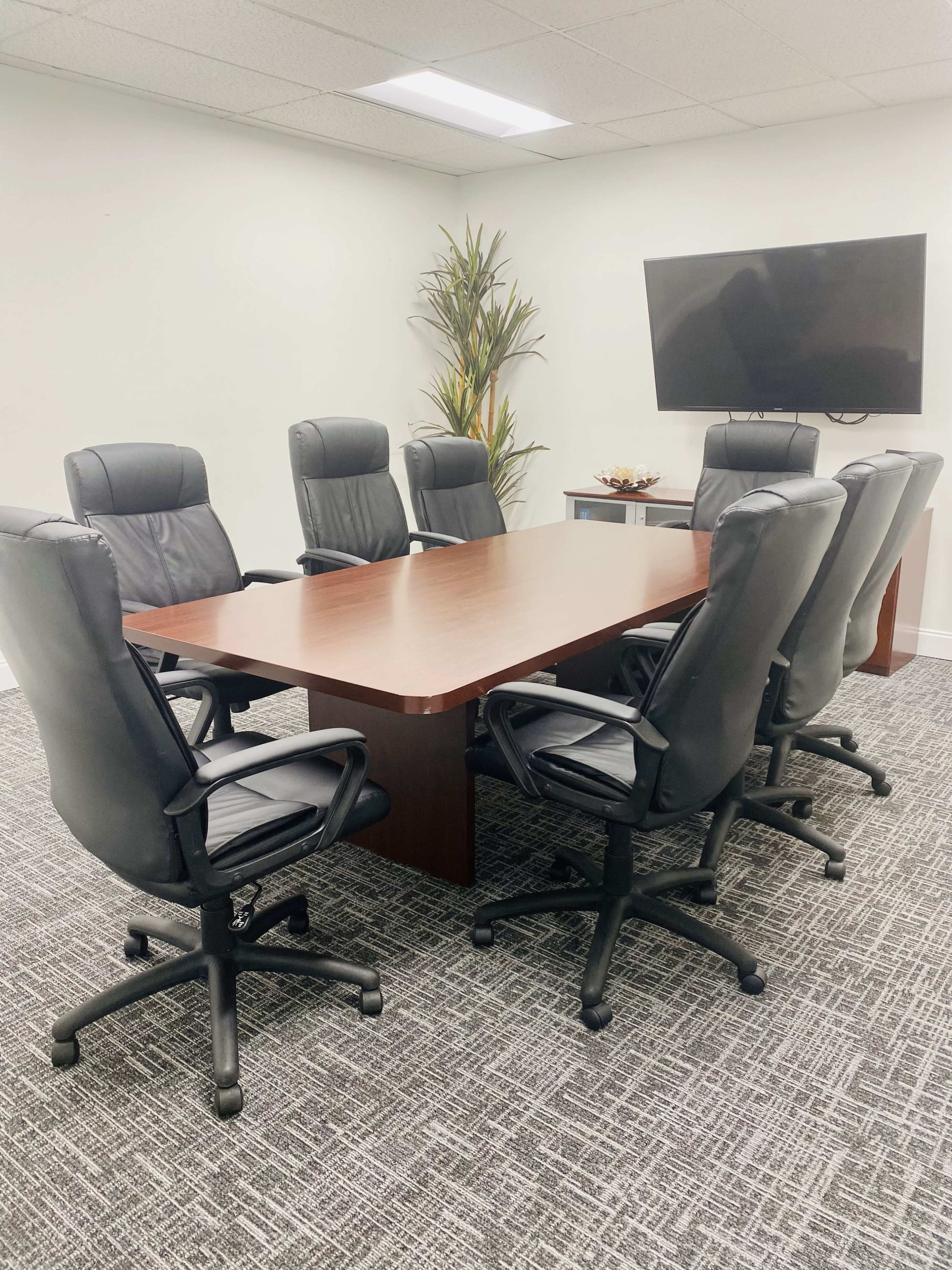 A conference room features a large wooden table surrounded by six black leather swivel chairs, with a plant and a wall-mounted television in the background.