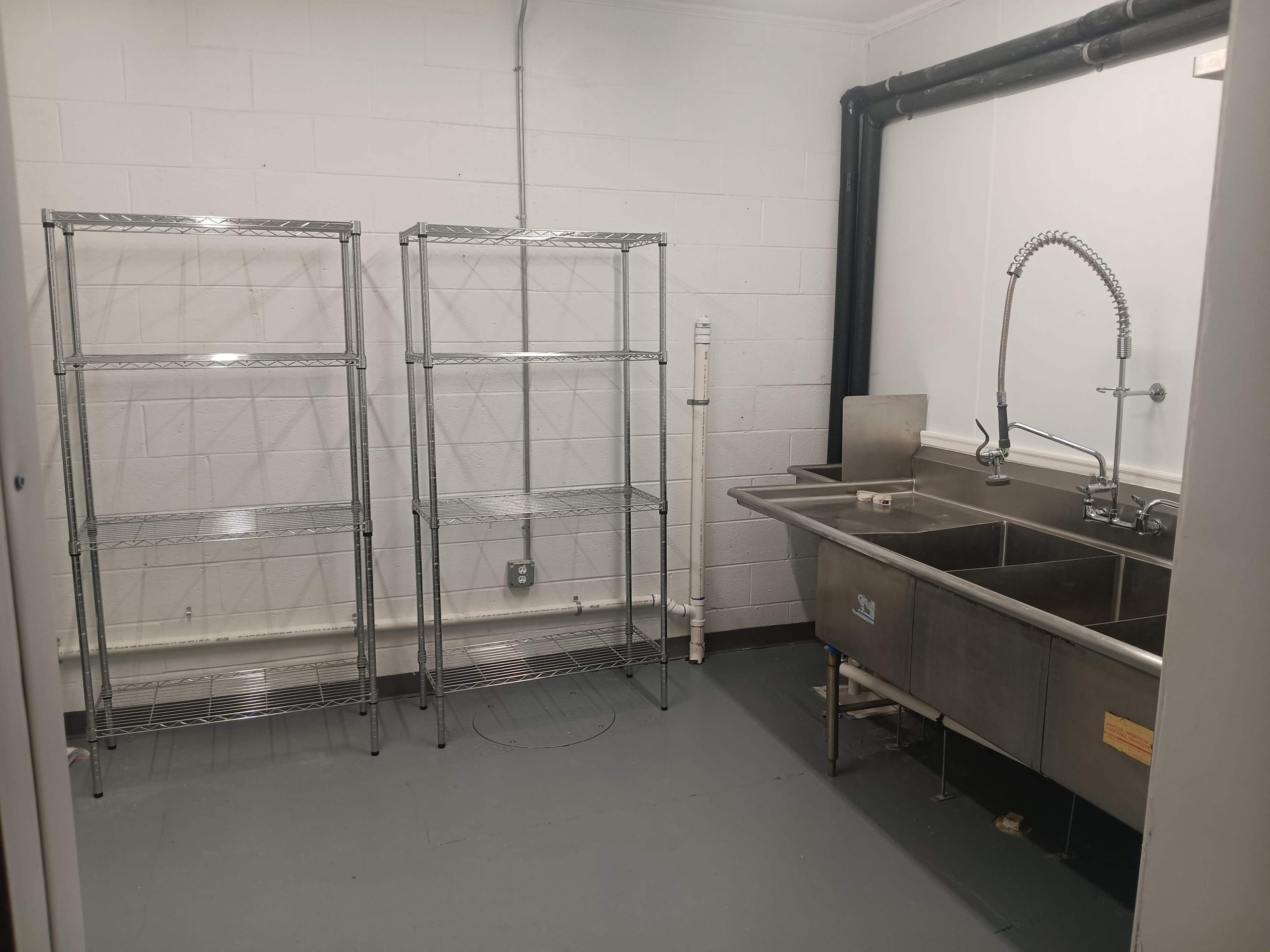 The image shows a small, bare room with two metal shelving units against one wall and a stainless steel sink with a faucet on the opposite side.