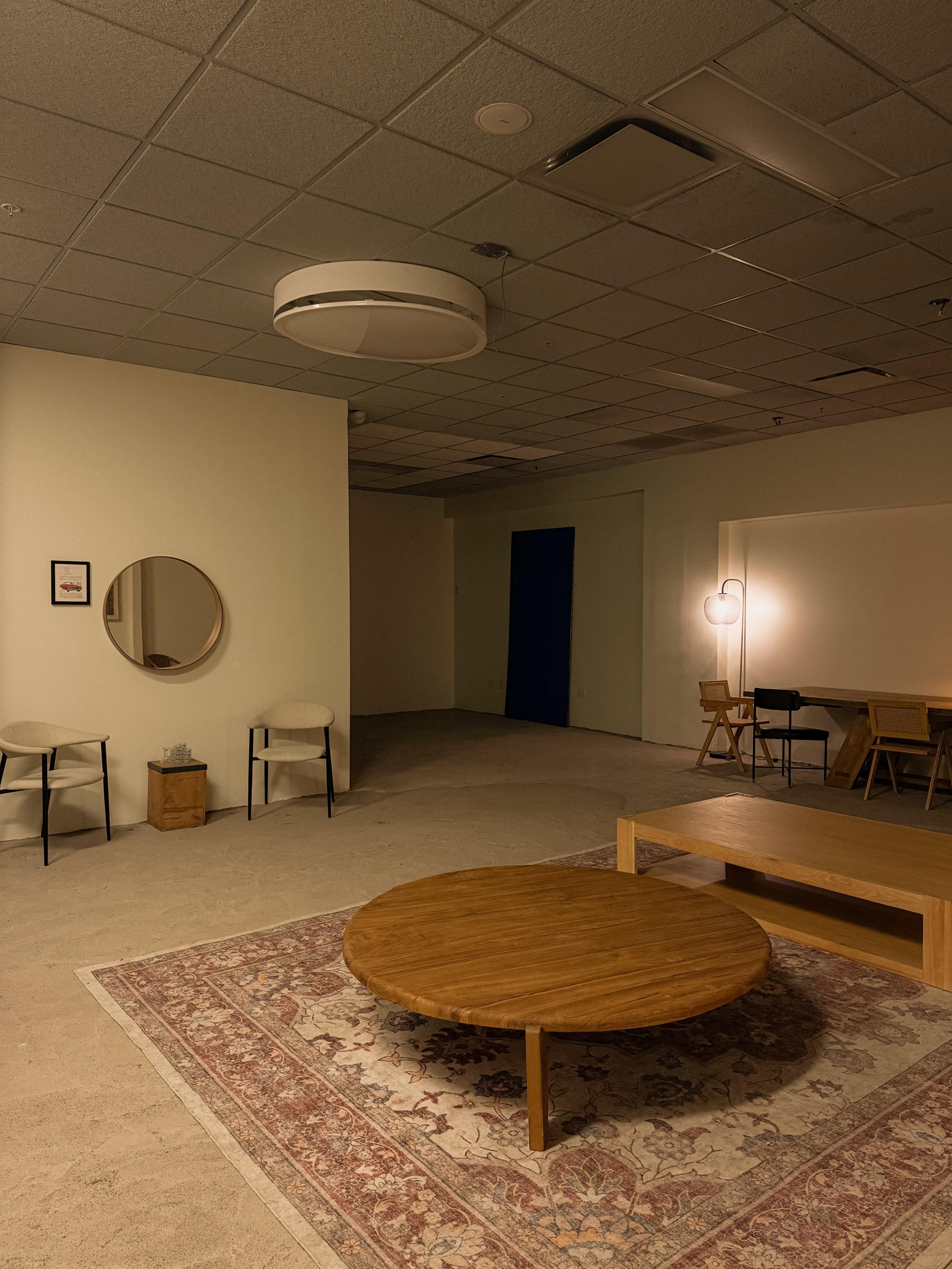 The image shows a spacious, dimly lit room featuring a circular wooden coffee table, a Persian rug, a round mirror, and various seating arrangements with a blue door in the background.