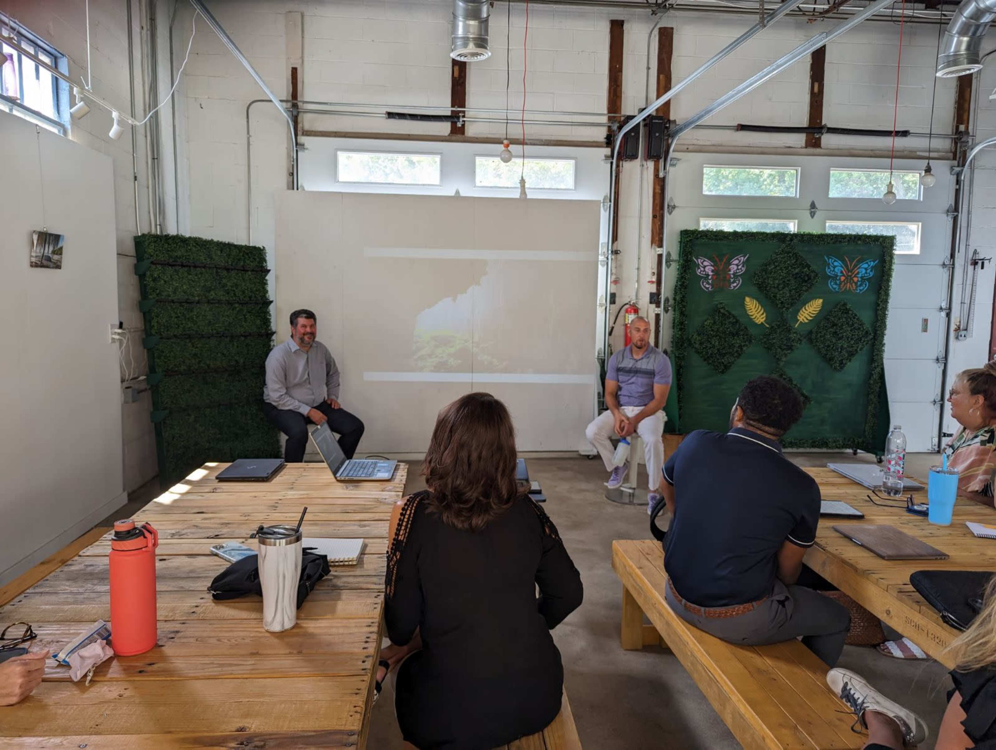 A group of people is seated around wooden tables in a spacious room, with two individuals at the front engaged in a discussion, and a projector displaying a nature scene on the wall.