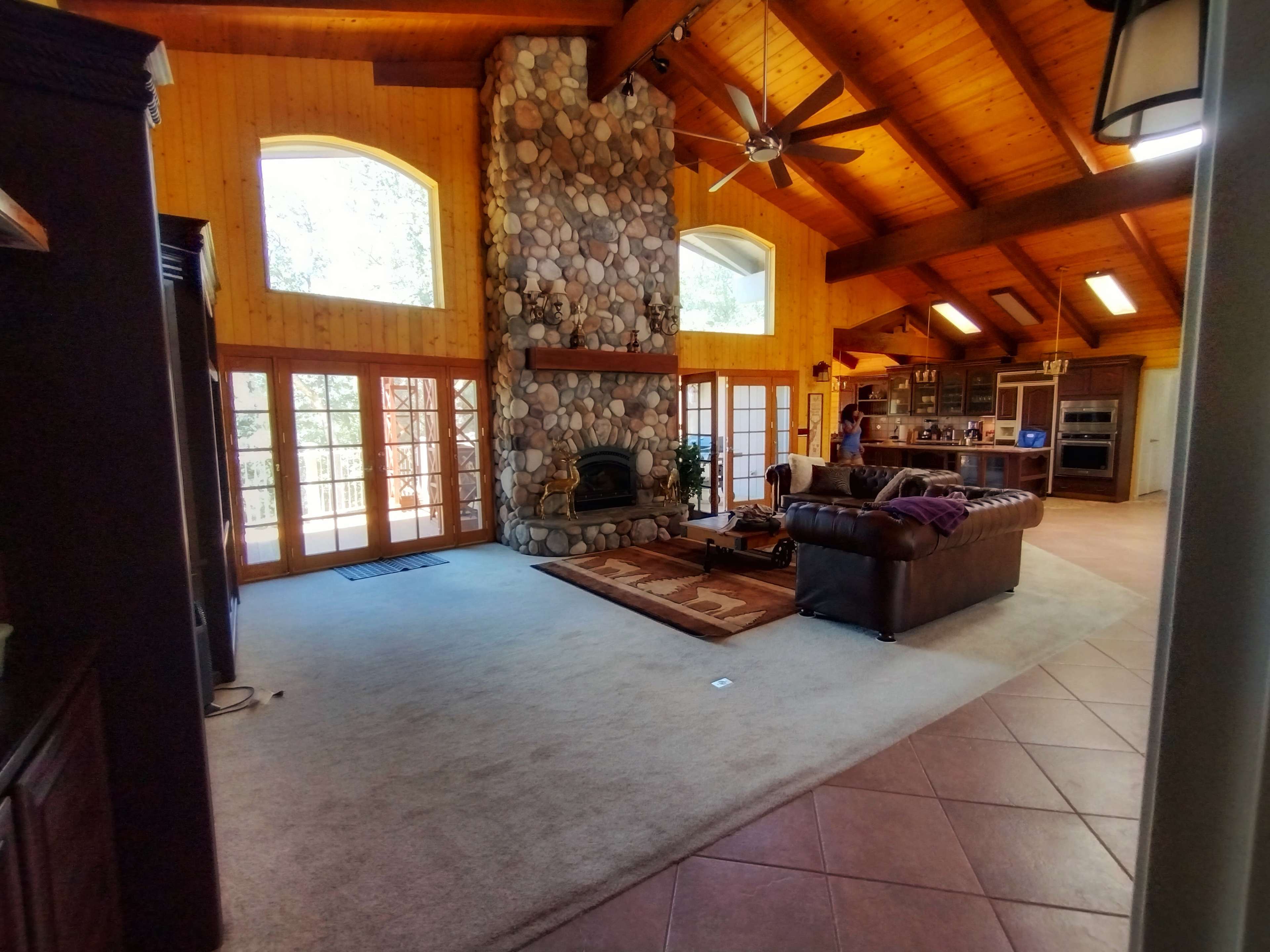 A spacious room features a stone fireplace, wooden beams, and large windows, with a couch and kitchen area visible in the background.