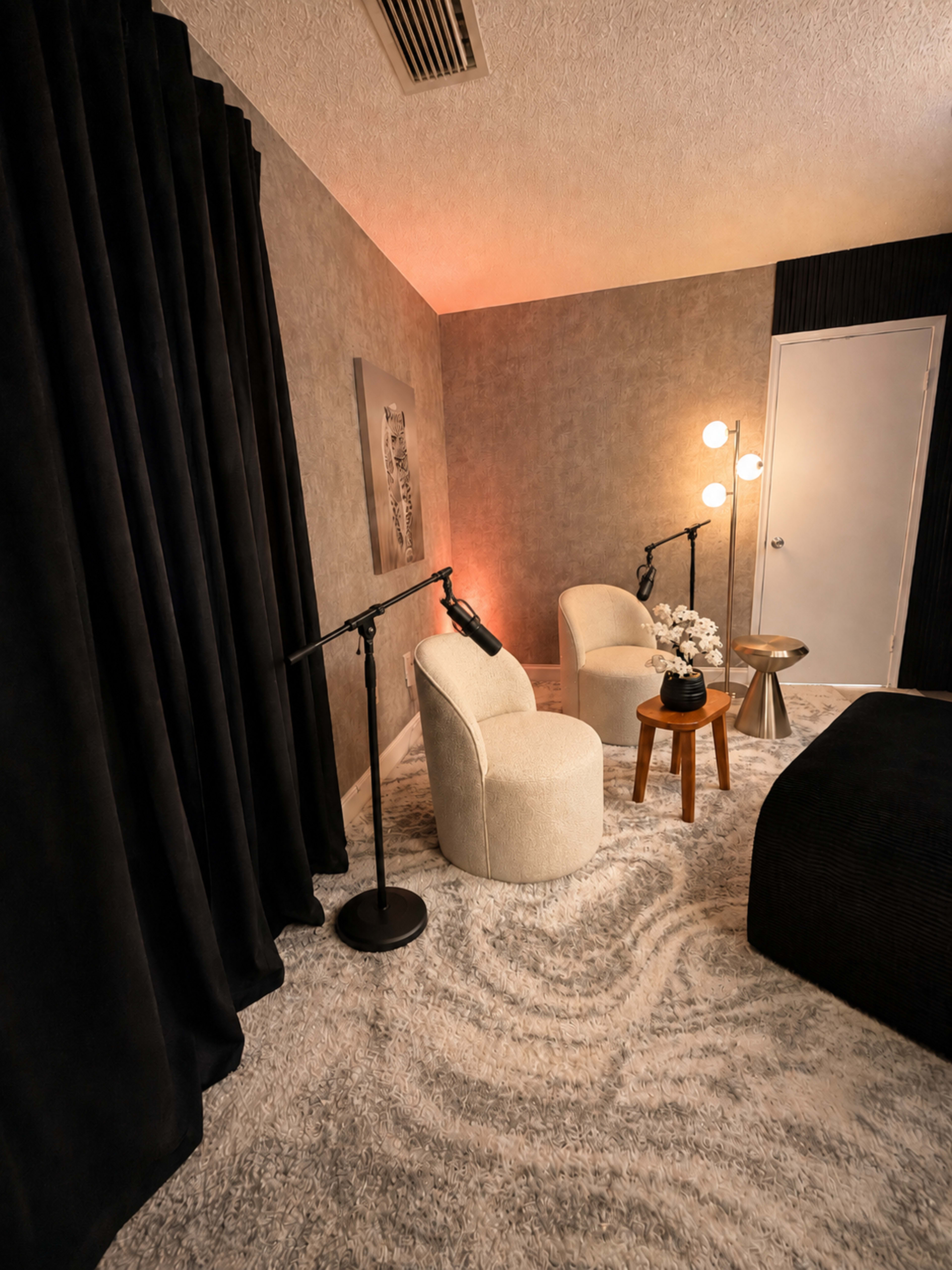 The image shows a cozy room featuring two upholstered chairs, a small side table, and a modern lamp, with dark curtains and textured walls creating a welcoming atmosphere.