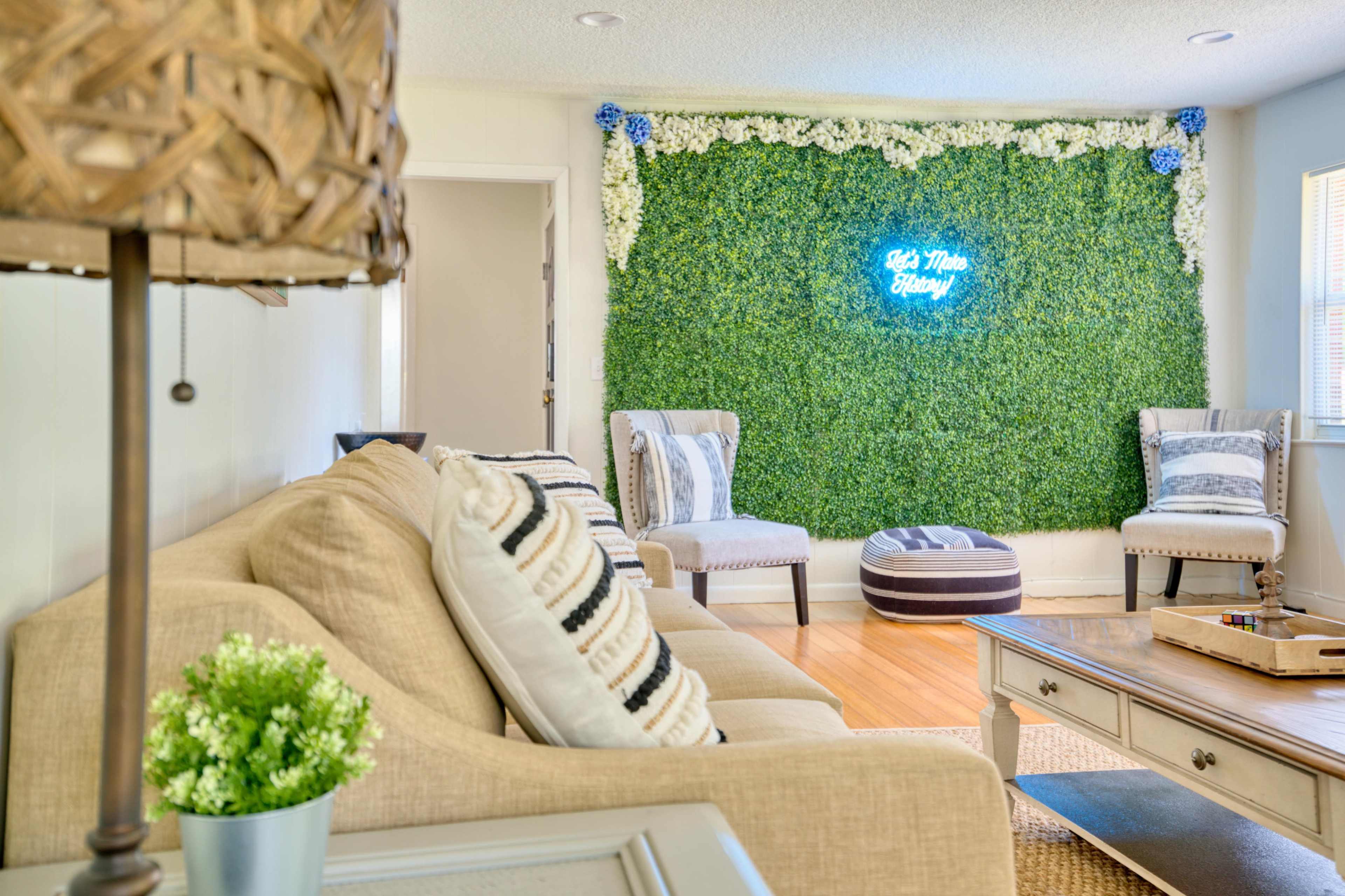 A living room features a beige sofa, two patterned chairs, a decorative coffee table, and a green wall adorned with white flowers and a glowing blue sign.