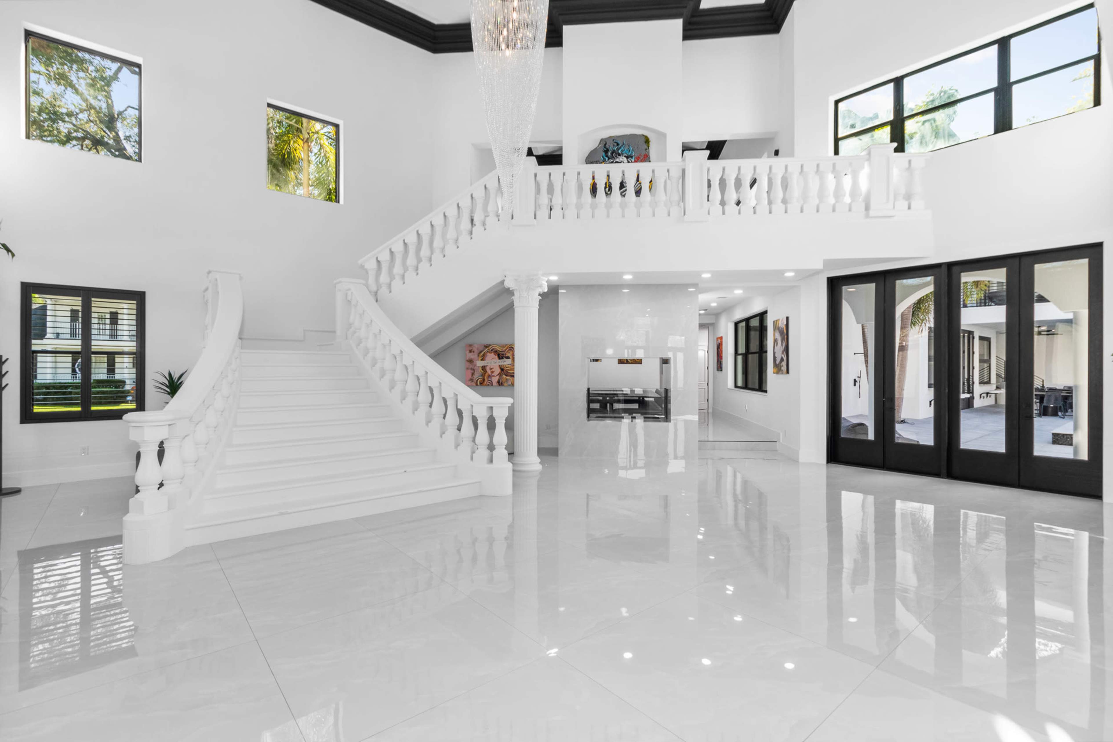 The image shows a spacious, modern foyer with a grand staircase, high ceilings, and large windows, featuring glossy white tile flooring.