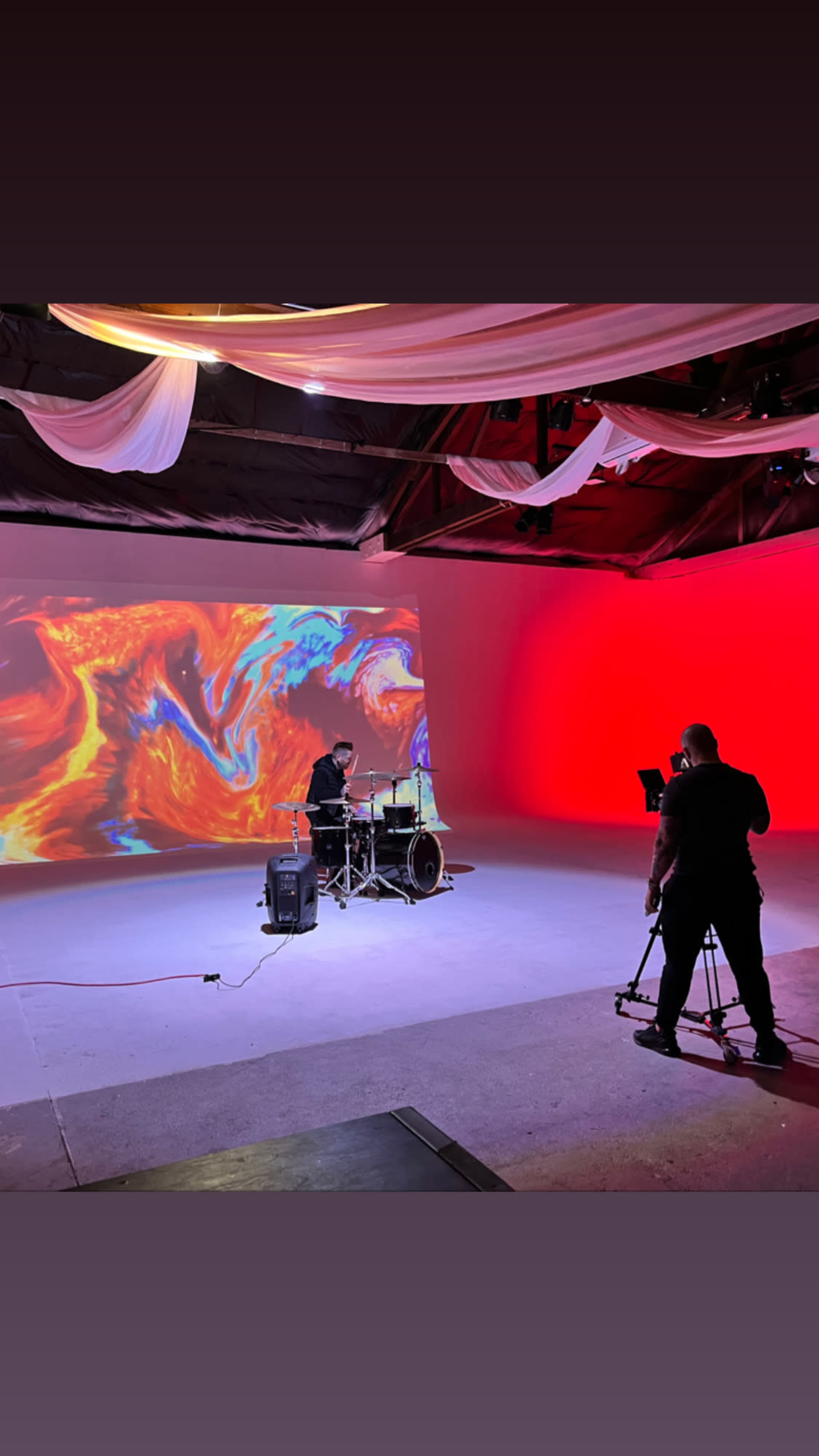 A drummer performs on stage in front of a large, colorful projected backdrop while a person filming captures the scene.